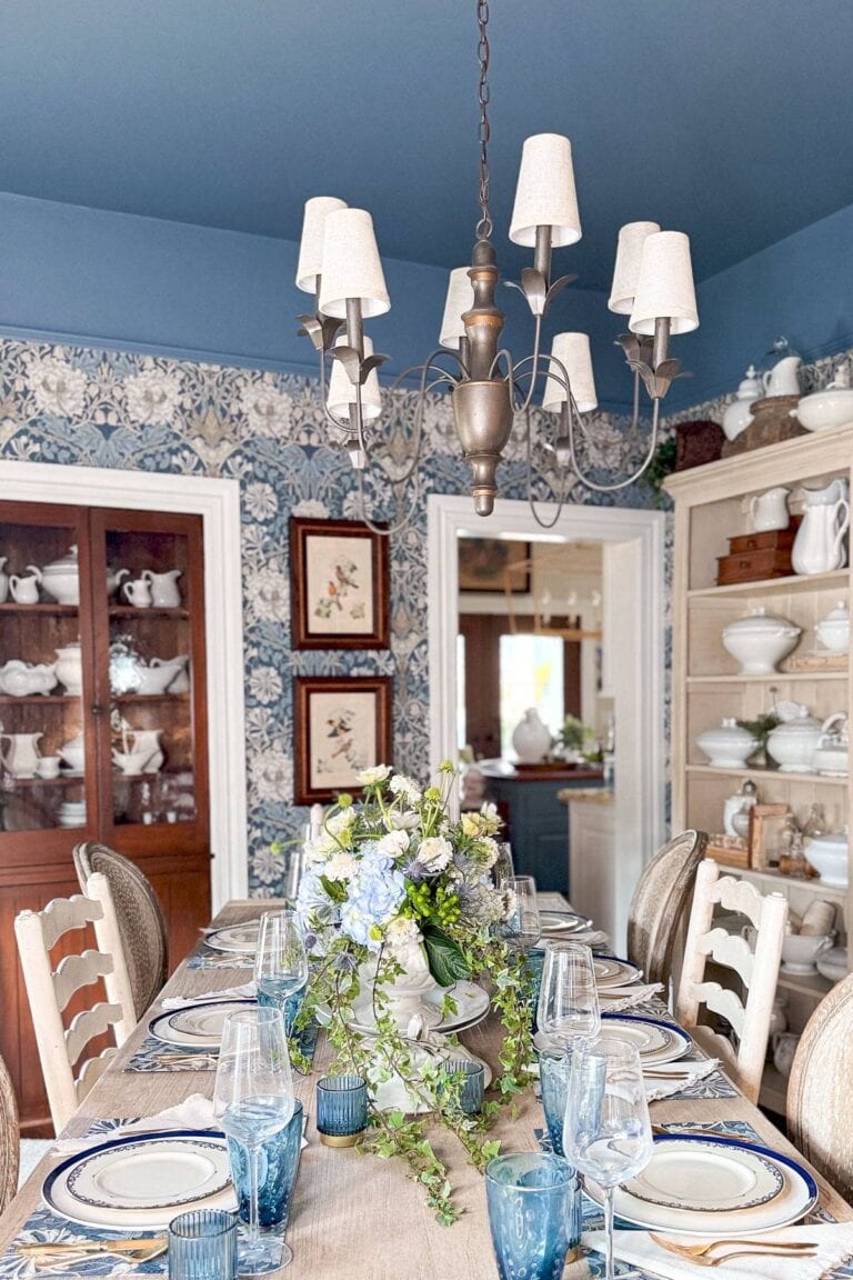 A dining room with a long table set for a meal. The table has blue and white plates, clear wine glasses, blue glass tumblers, and a floral centerpiece. A chandelier hangs above. The room has blue wallpaper and shelves with white dishes.