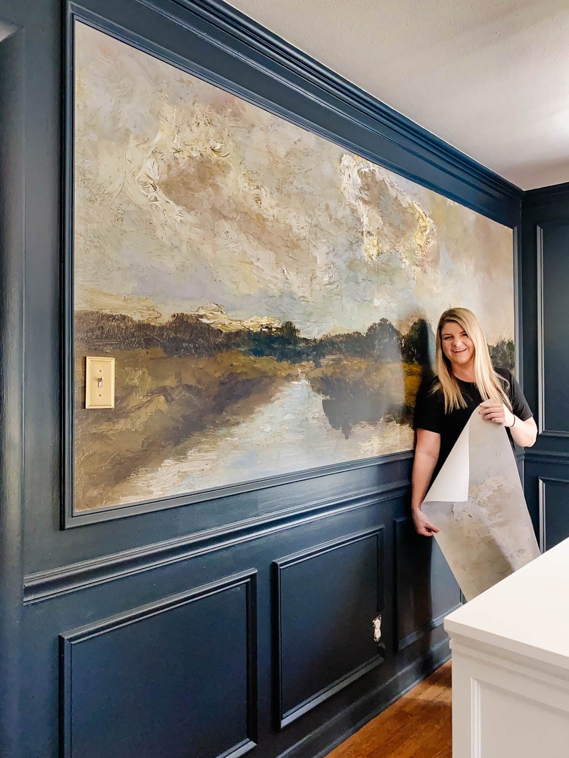 A woman stands smiling in front of a large, textured landscape painting on a dark-paneled dining room wall. She holds a rolled section of wallpaper that matches the painting's scenery, perfectly updating the aesthetic. A light switch is visible on the wall.