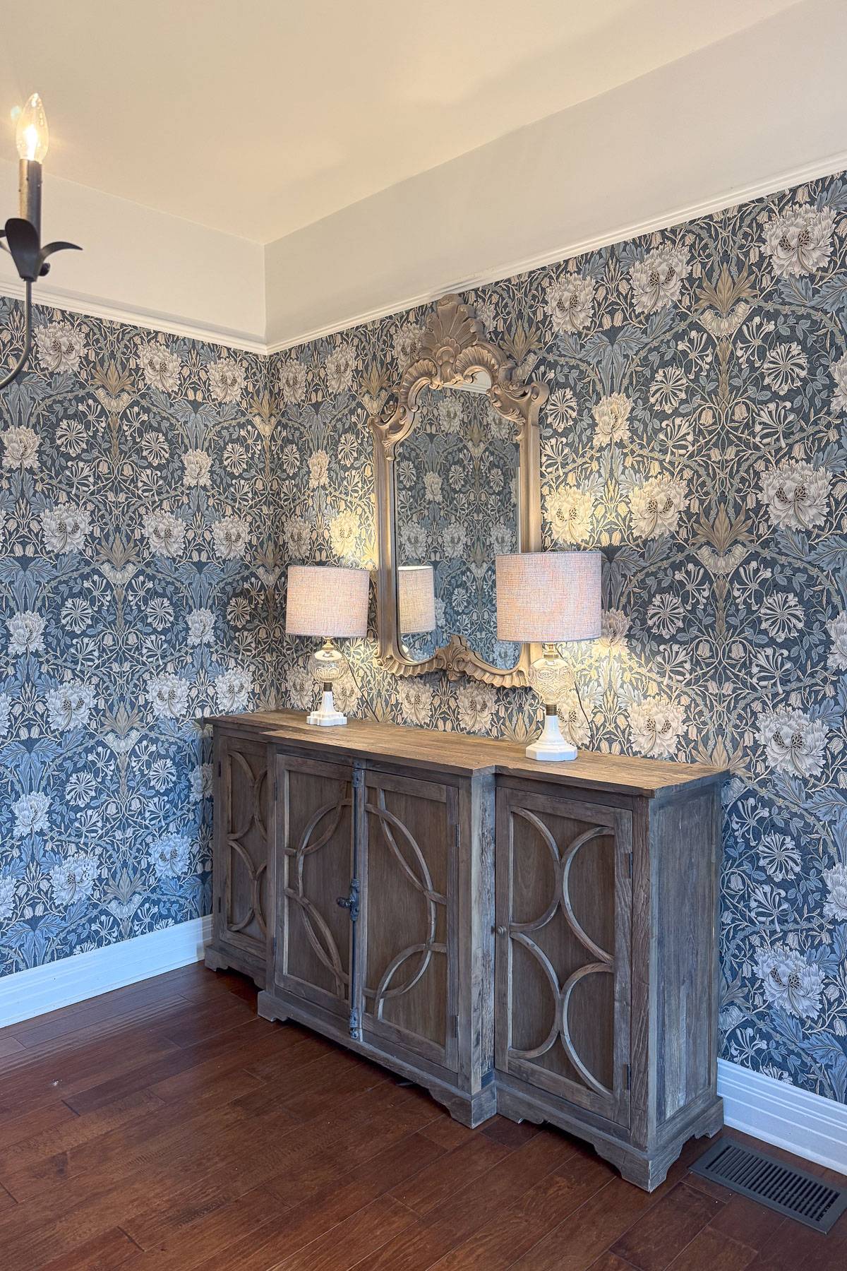 A room features a wooden sideboard with decorative circular patterns, topped with two lamps. Above it hangs an ornate mirror. The walls are adorned with floral-patterned wallpaper in blue and white tones. The floor is wooden, and a chandelier is partially visible.