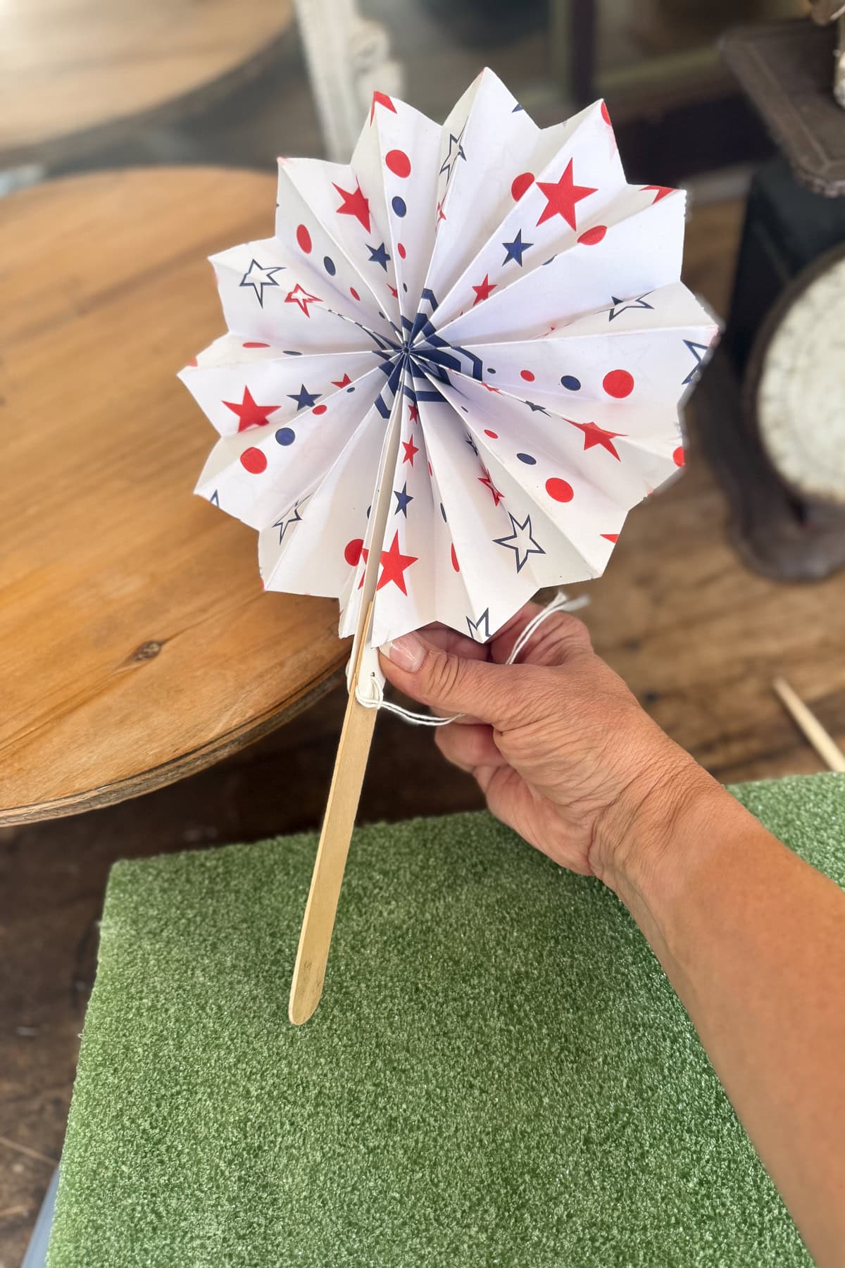 A hand holds a paper fan decorated with red and blue stars and dots. The fan has a wooden handle and is set against a background of a wooden table and green textured surface.