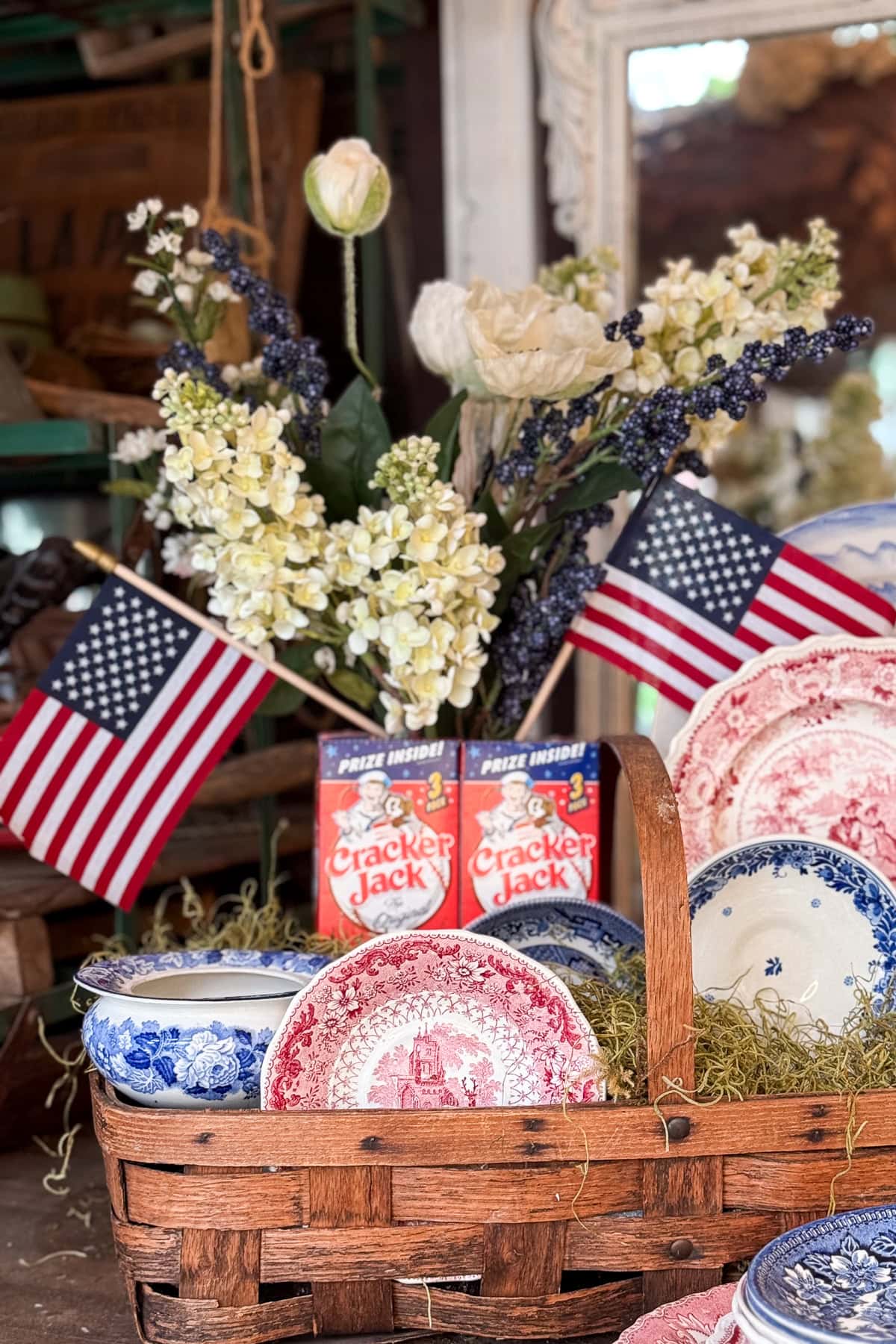 A basket of blue and red vintage dishes sits on straw, with two boxes of Cracker Jack, American flags, and a bouquet of white and purple flowers in the background.