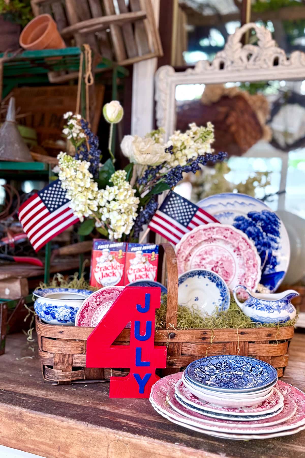 A rustic table display features vintage red, white, and blue dishes, small American flags, Cracker Jack boxes, and a large red "4 JULY" sign, with flowers in the background, evoking a patriotic 4th of July theme.