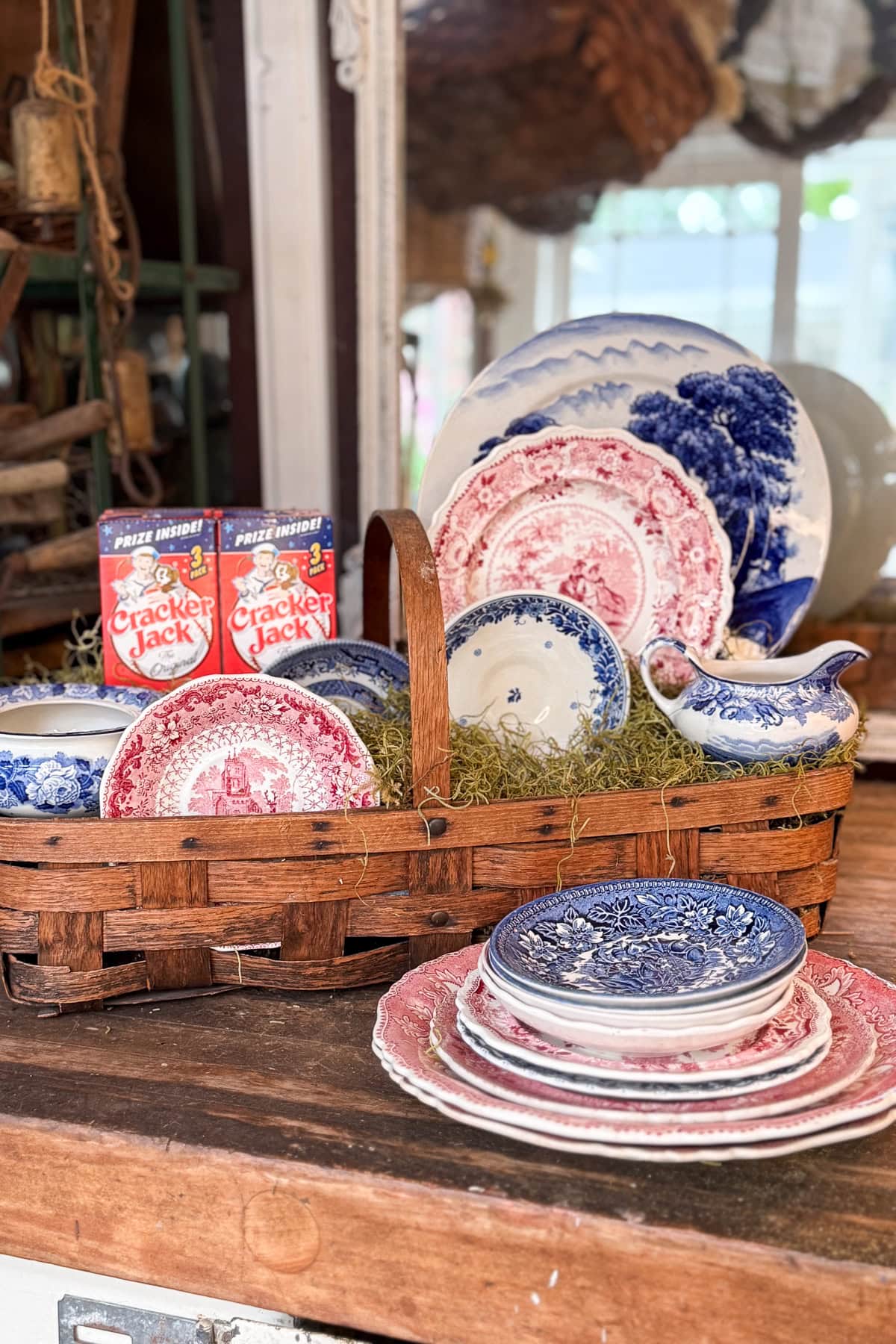 A wicker basket filled with vintage blue and red patterned plates and bowls sits on a wooden table, with two boxes of Cracker Jack popcorn in the background and more stacked dishes nearby.