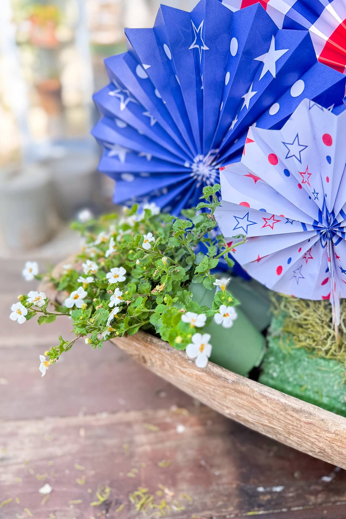 A wooden bowl holds small white flowers and decorative paper fans in blue and white with red, blue, and white star and dot patterns, resting on a rustic wooden surface.