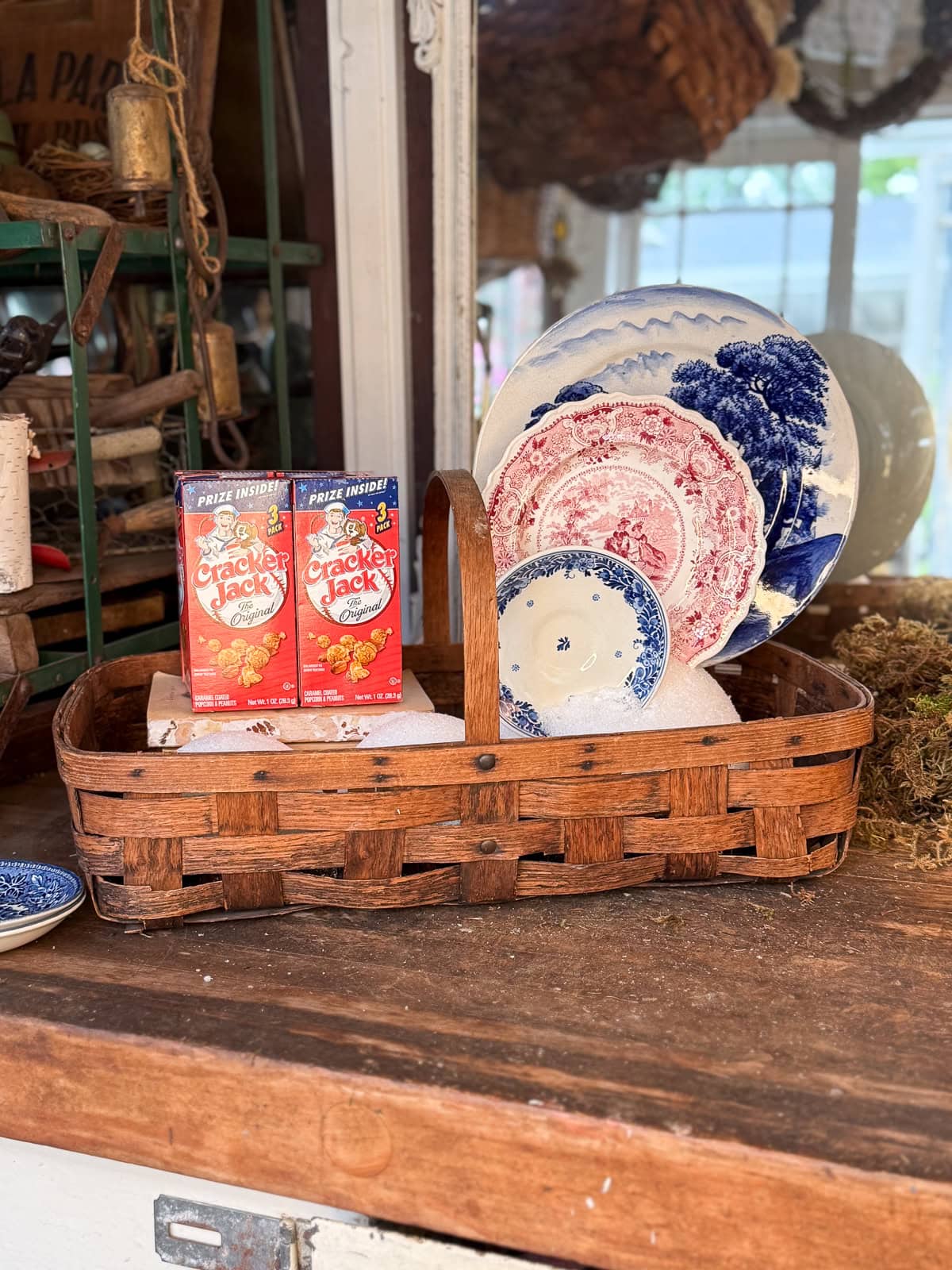 A woven basket holds vintage patterned plates in blue and red alongside two boxes of Cracker Jack on a wooden surface, with rustic decor and shelves in the background.