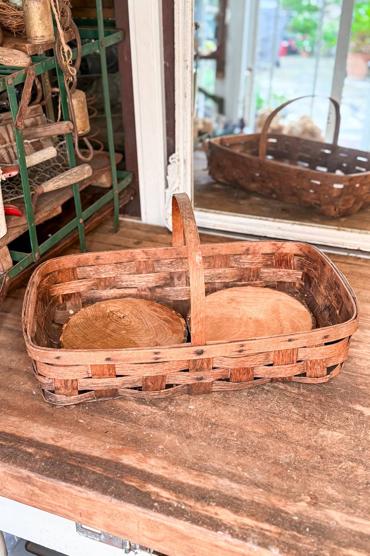 A rectangular woven basket with a handle sits on a wooden surface. Inside the basket are two round wooden slices. In the background, there is a window and various objects on shelves.