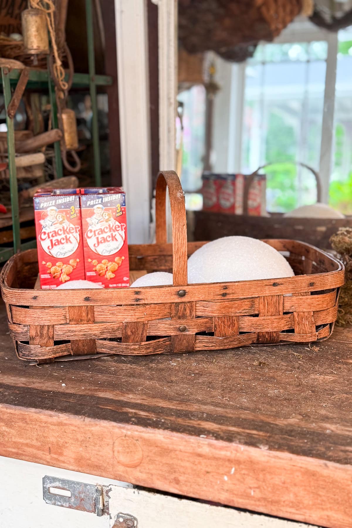 A woven wooden basket on a wooden counter holds two boxes of Cracker Jack and some white round objects. The background shows a window with a blurry outdoor view and shelves with rustic decor.