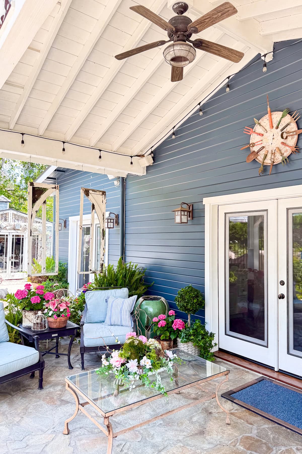A cozy patio with blue siding, white French doors, and a vaulted white ceiling with a fan. There are potted flowers, cushioned seating, a glass coffee table, and string lights creating a welcoming outdoor space.
