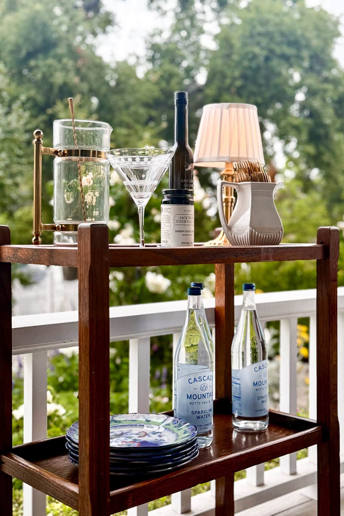 A wooden bar cart on a porch holds cocktail glasses, a carafe, plates, and sparkling water—an effortless touch for easy porch decor. Green trees in the background complete this inviting summer porch idea.