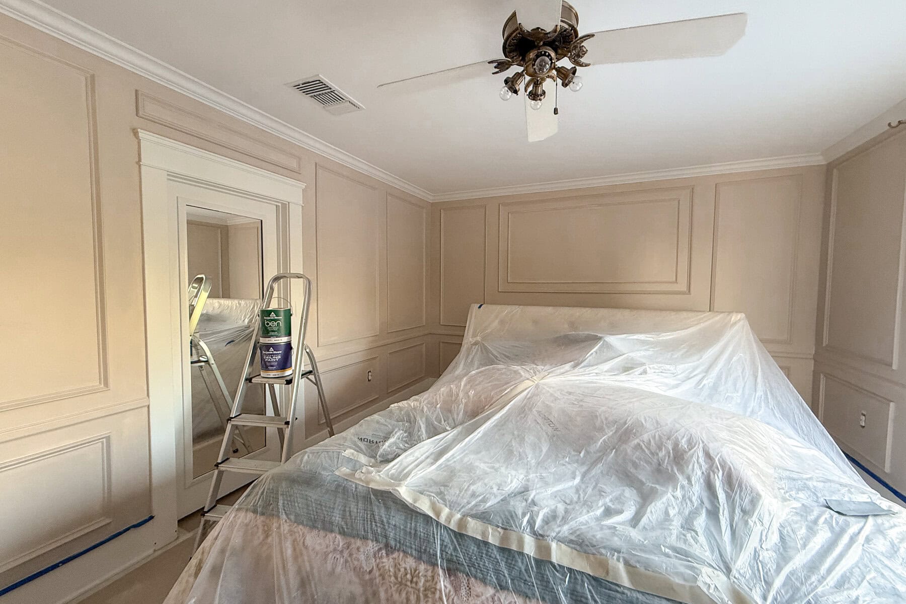 A bedroom prepared for painting, with a bed covered in plastic sheeting, a ladder holding a paint can and supplies, and light beige paneled walls ready for DIY wall moldings. A ceiling fan and a large mirror are also visible.