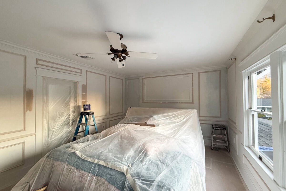A room being painted, with walls partially painted beige, furniture covered in plastic, a step ladder, paint cans, and sunlight coming through the windows. A ceiling fan hangs in the center of the ceiling.