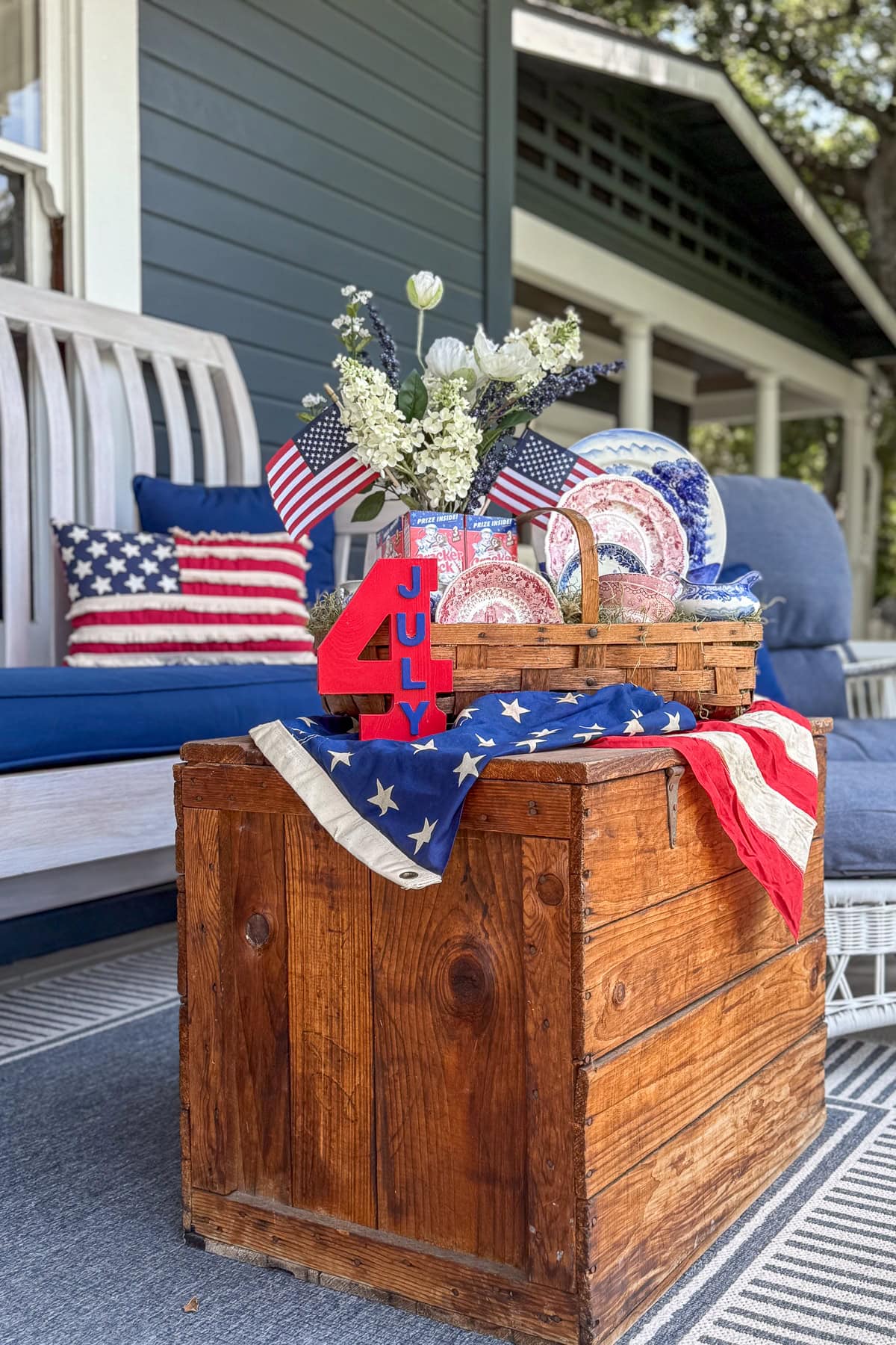 A rustic wooden table decorated with an American flag, red, white, and blue tableware, small U.S. flags, flowers, and a red "4th of July" sign, set on a porch with patriotic pillows and blue seating.