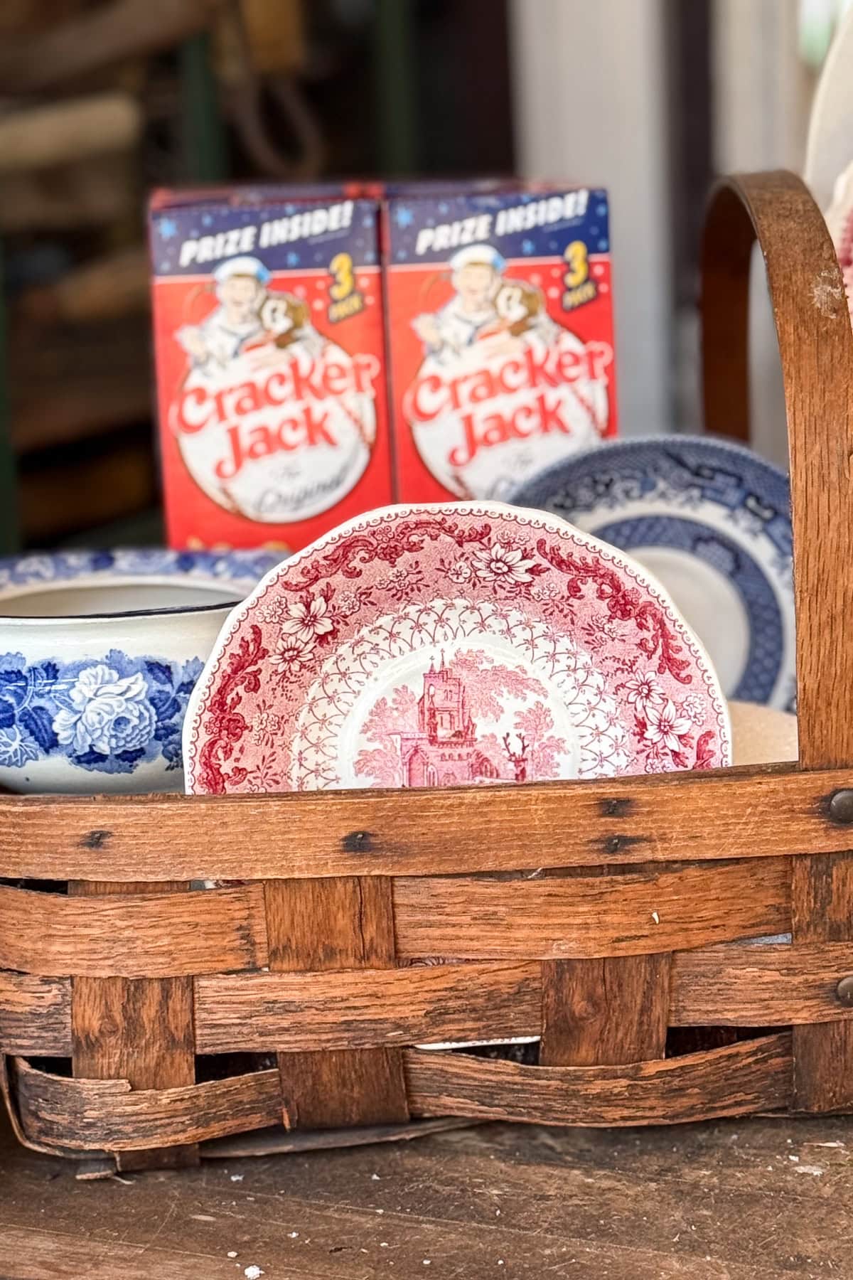 A woven wooden basket holds vintage blue and red patterned plates, with two Cracker Jack boxes standing behind them in the background.