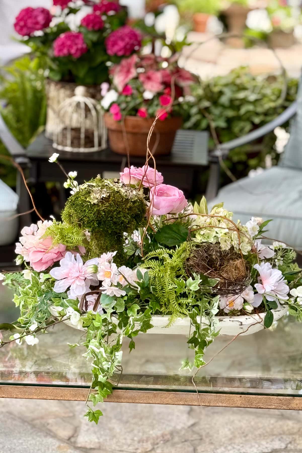 A floral centerpiece with pink roses, moss, ferns, and trailing greenery sits on a glass table. In the background, potted flowers, a birdcage, and patio chairs are visible in an outdoor garden setting.