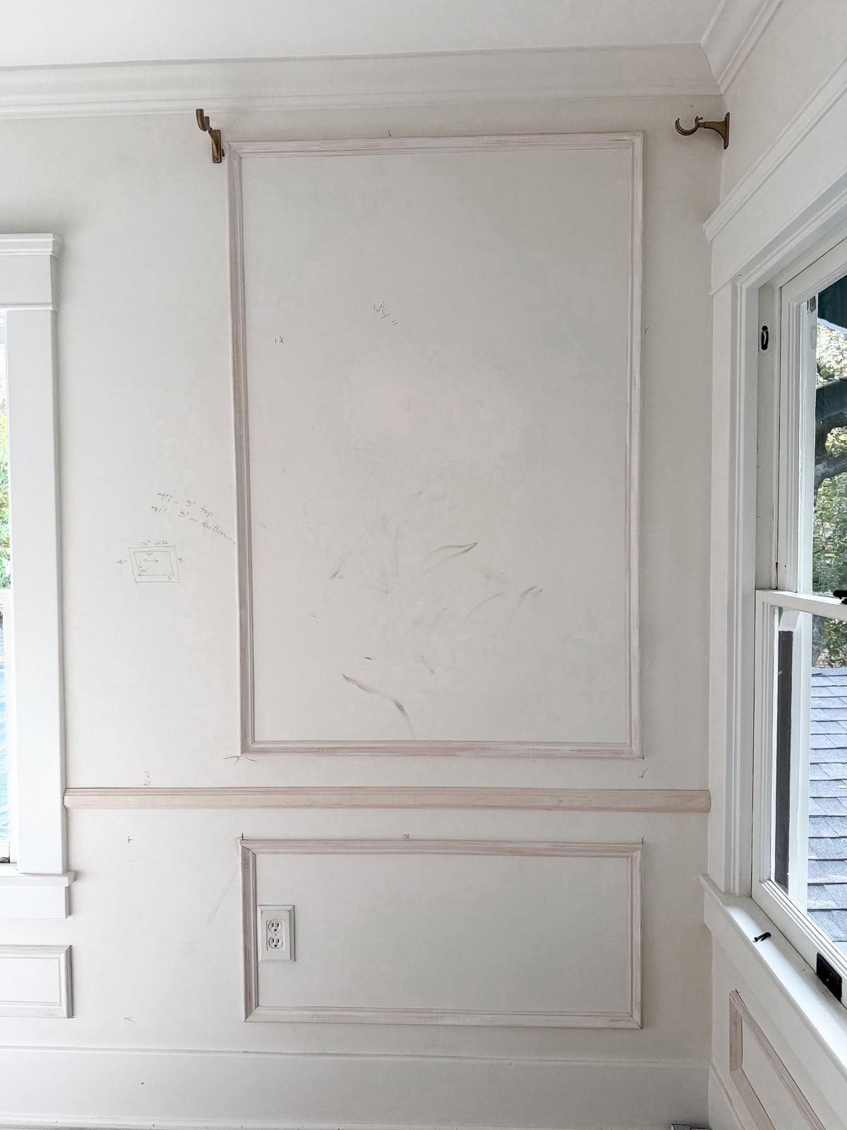 A white wall with DIY wall moldings, scuff marks, a visible electrical outlet, and brass curtain rod brackets near the ceiling. There is a window on the right letting in natural light.