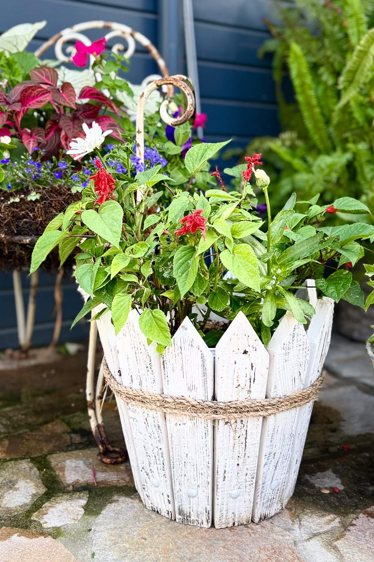 A white, rustic wooden planter filled with green leafy plants and red flowers sits on a stone patio. A metal chair with colorful flowers and a fern are in the background, creating a cozy garden scene.