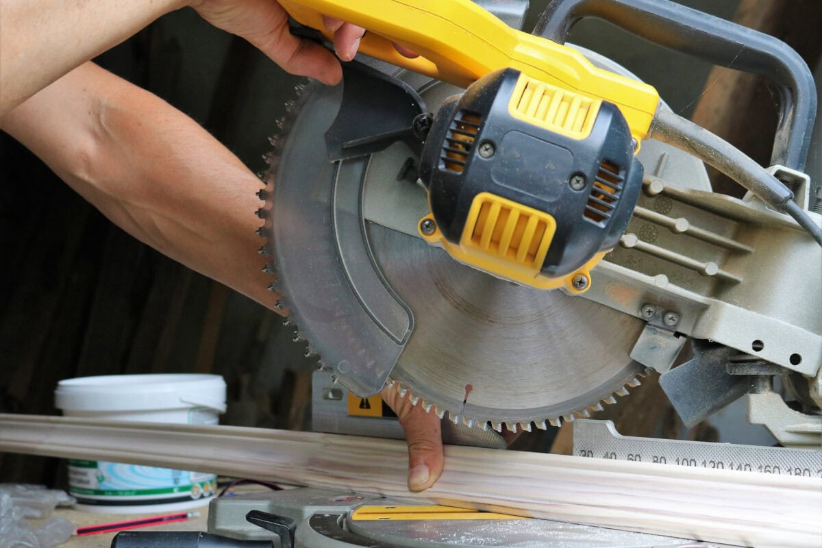A person uses a circular miter saw to cut wooden planks, with their hand holding the wood close to the sharp spinning blade, demonstrating a potentially unsafe practice.