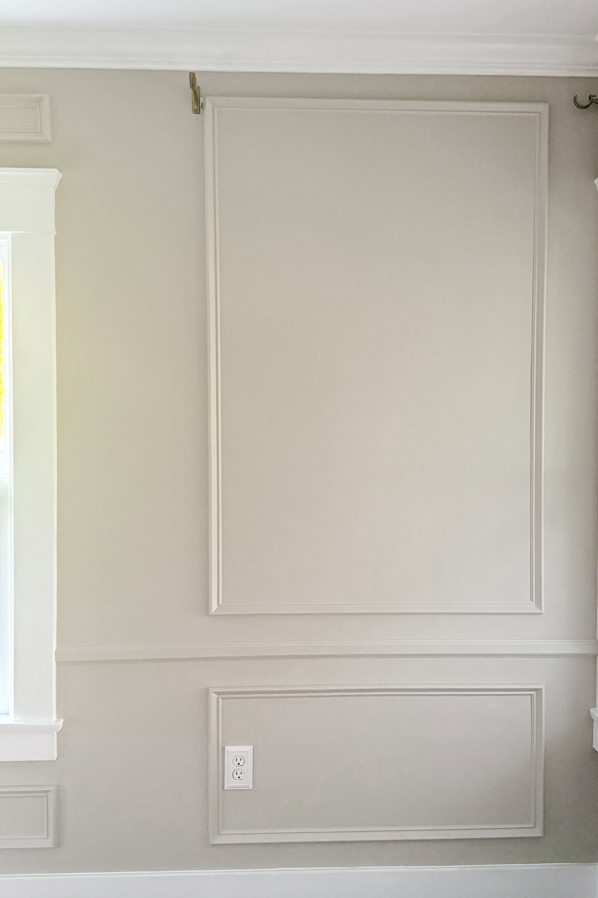 A light gray wall with two rectangular decorative trim panels, one large above and one small below. A white electrical outlet is on the lower panel, and part of a window frame is visible on the left side.