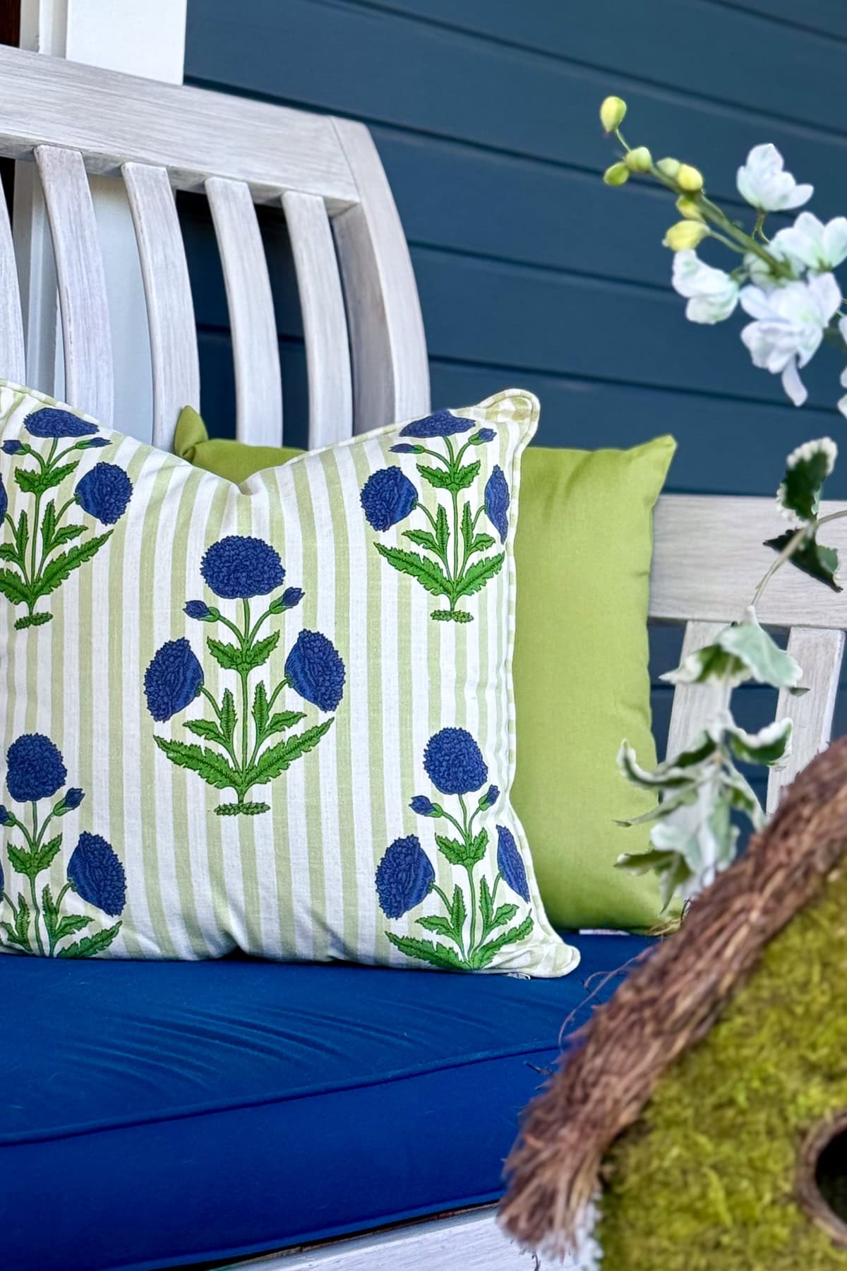 A white wooden bench with a blue seat cushion, green and white striped pillow with blue floral embroidery, a plain green pillow, and white flowers in the background. A mossy decorative object is in the foreground.
