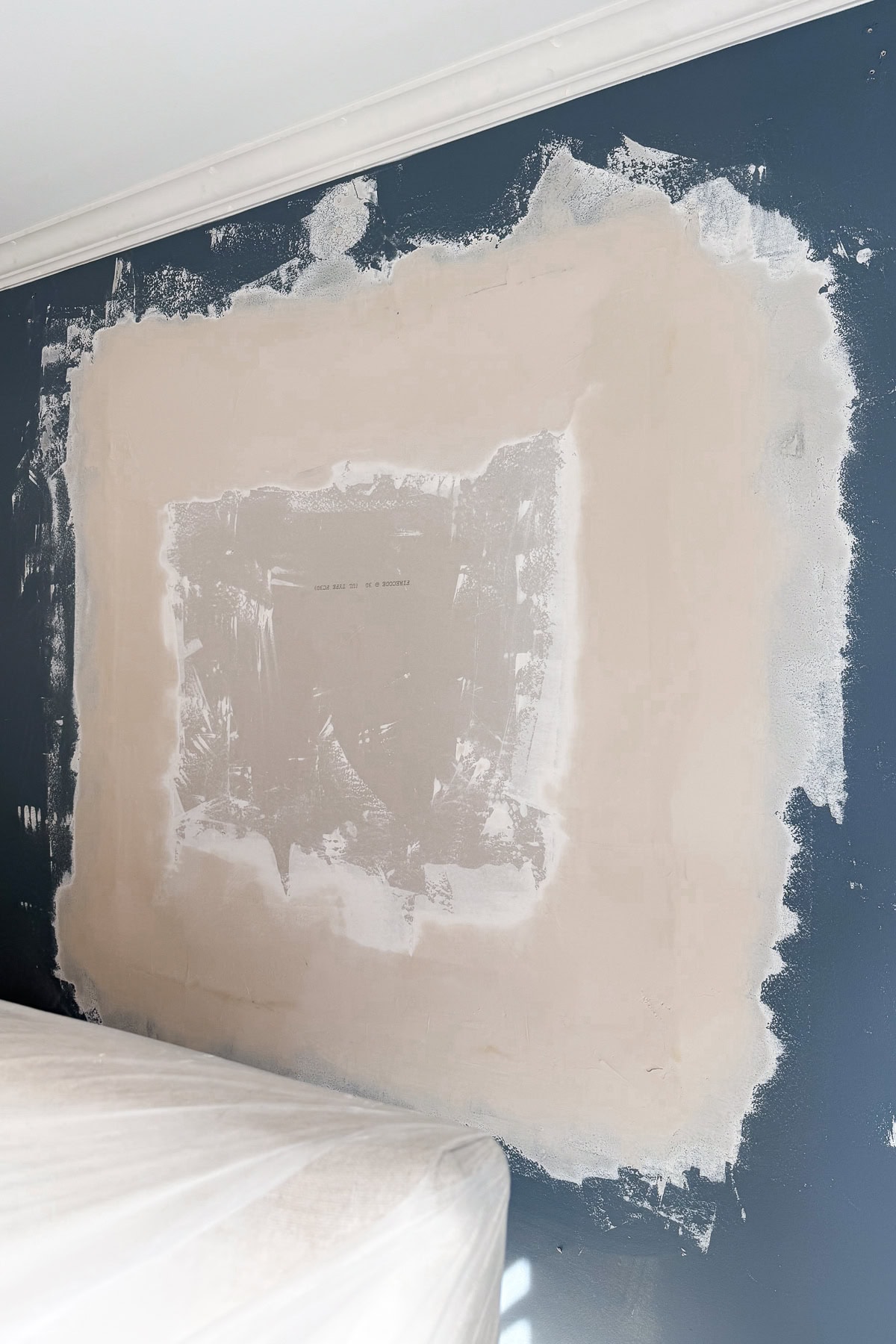 A wall with blue paint has a large, roughly rectangular patch of white spackle in the center, indicating recent repairs. The area around the patch is uneven, and a covered piece of furniture is partially visible at the bottom.