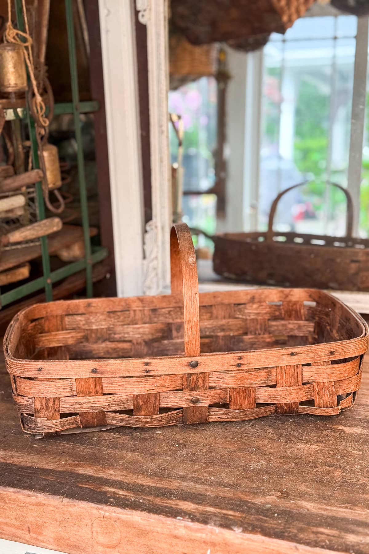 A rectangular woven wooden basket with a single handle sits on a rustic wooden surface in a cozy, vintage setting.