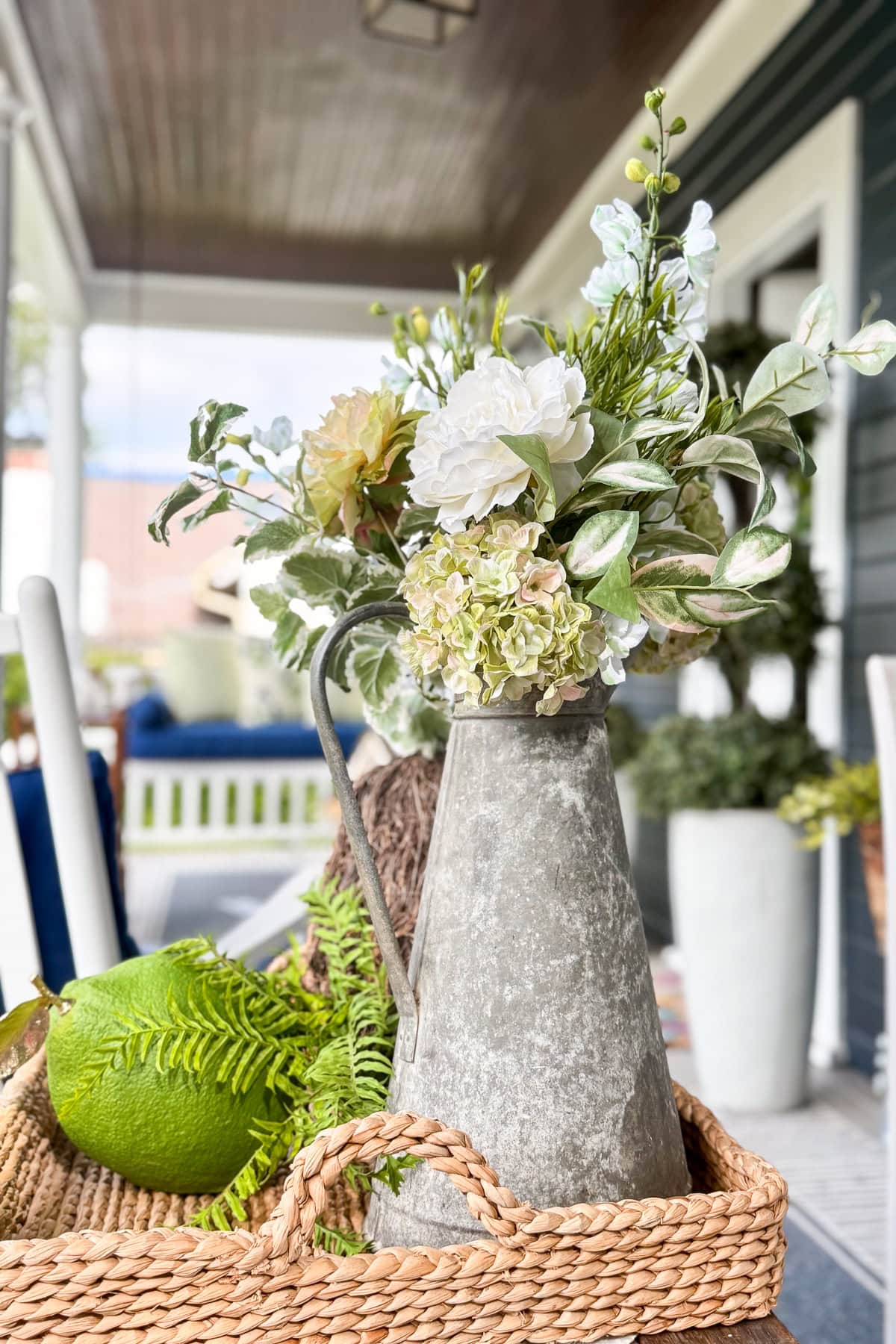 A rustic metal pitcher filled with white and green flowers sits in a woven basket on a porch, with a green decorative ball and a fern next to it, and outdoor furniture in the background.