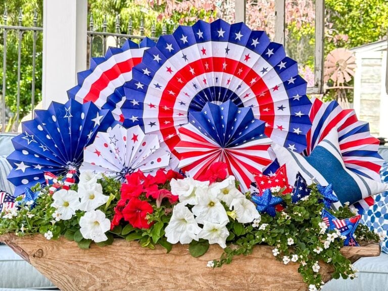 A festive arrangement with red, white, and blue paper fans, stars, and American flag patterns, surrounded by white and red flowers in a wooden container, celebrating a patriotic occasion.