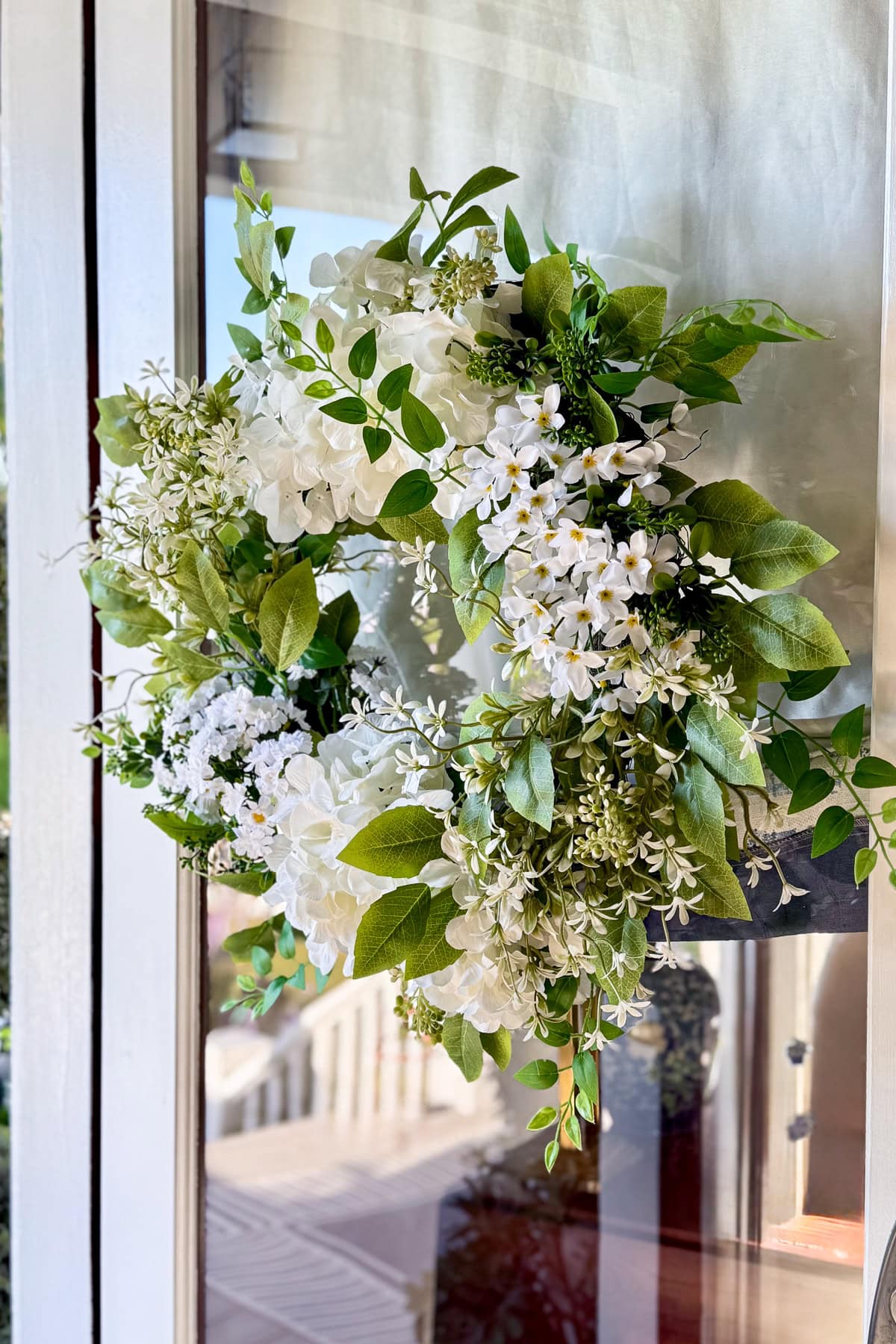 A wreath made of white flowers and green leaves hangs on a glass door, with sunlight shining through—a perfect touch for easy porch decor and charming summer porch decorations.