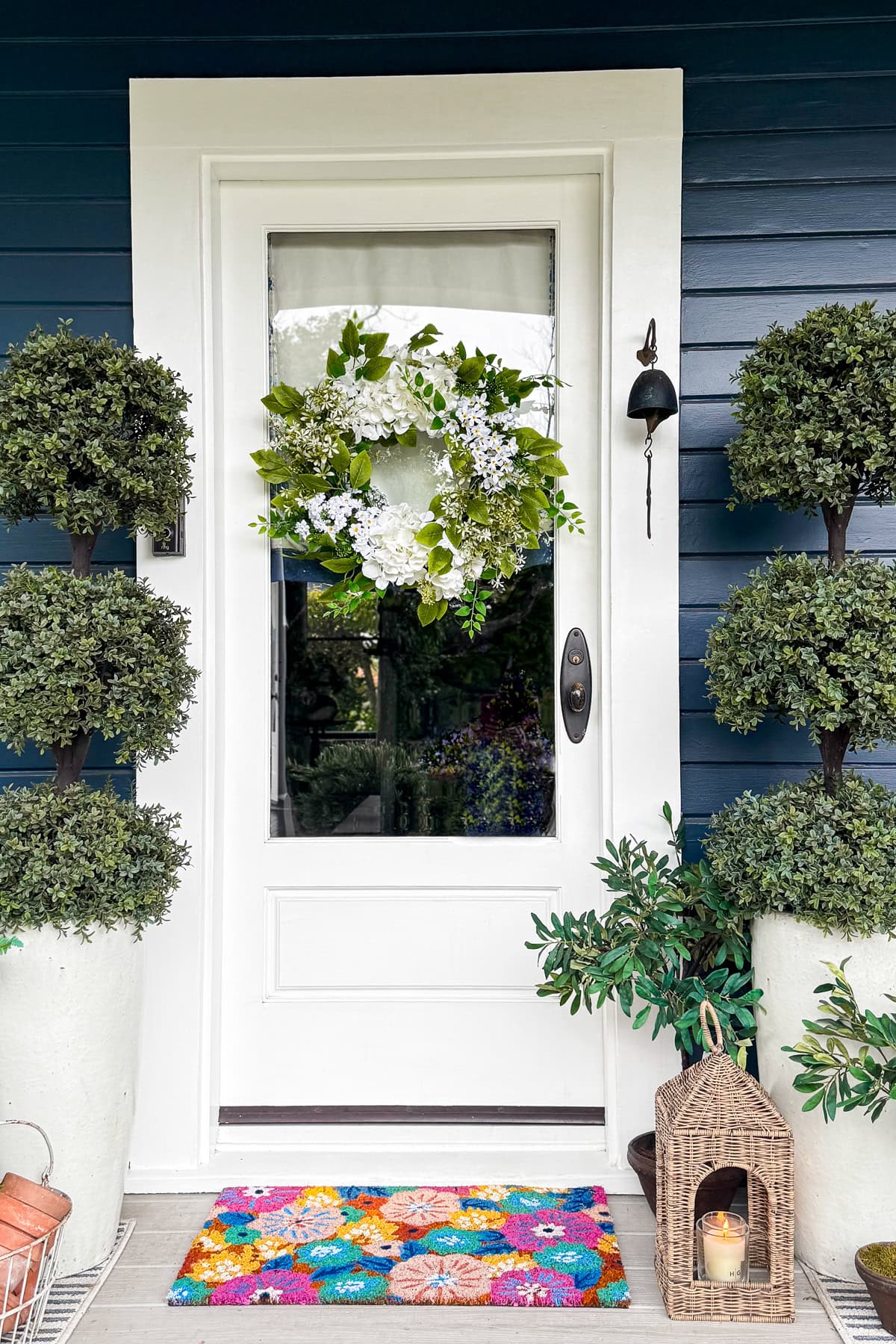 A white front door with a glass pane features a green and white floral wreath. Flanked by potted topiary trees, a colorful floral doormat, and a lantern with a candle sit on the blue-paneled porch.