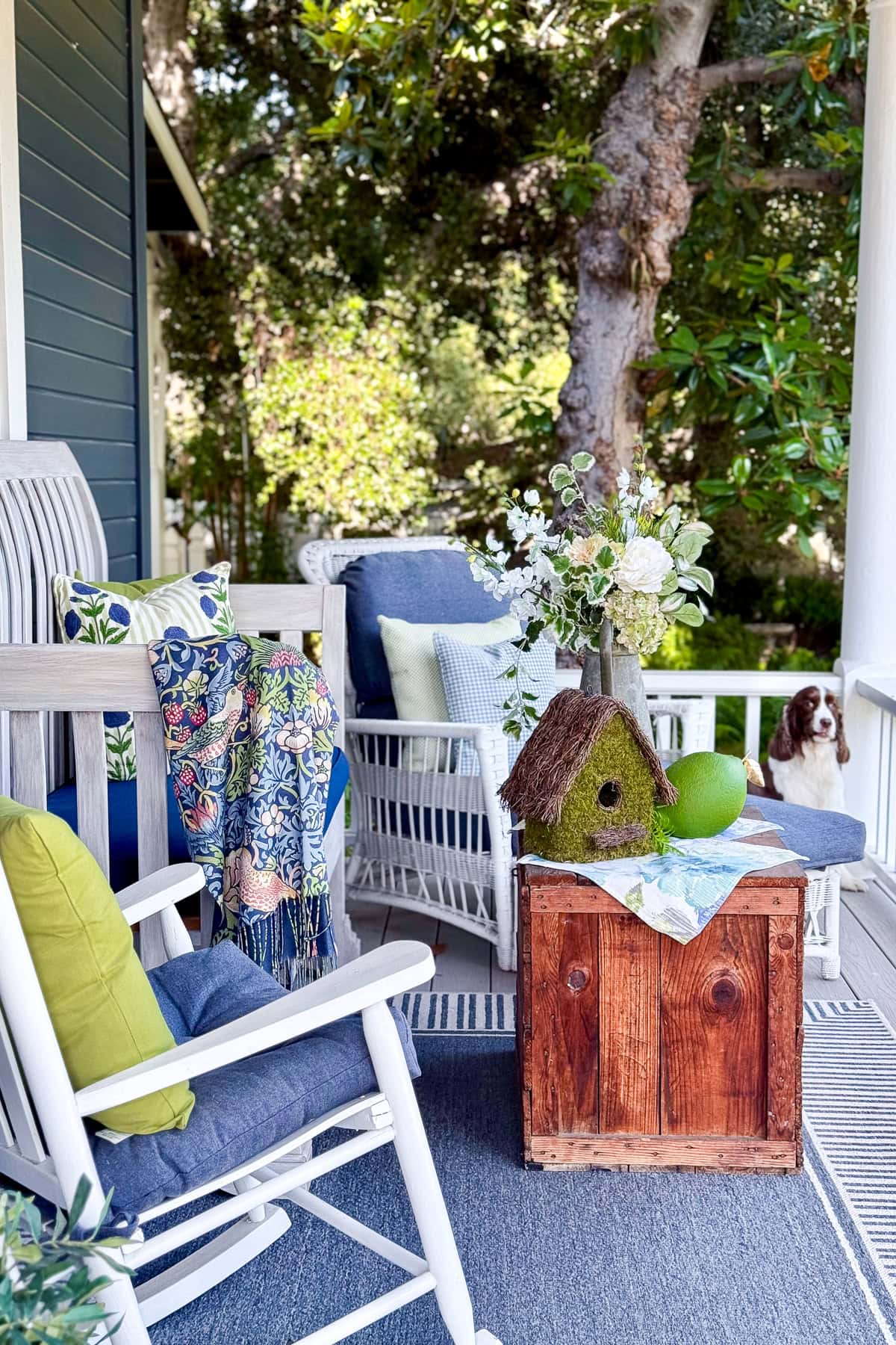 A cozy porch with white rocking chairs, colorful pillows and blanket, a wooden table with a mossy birdhouse, green apples, flowers, and a dog sitting in the background near lush trees.