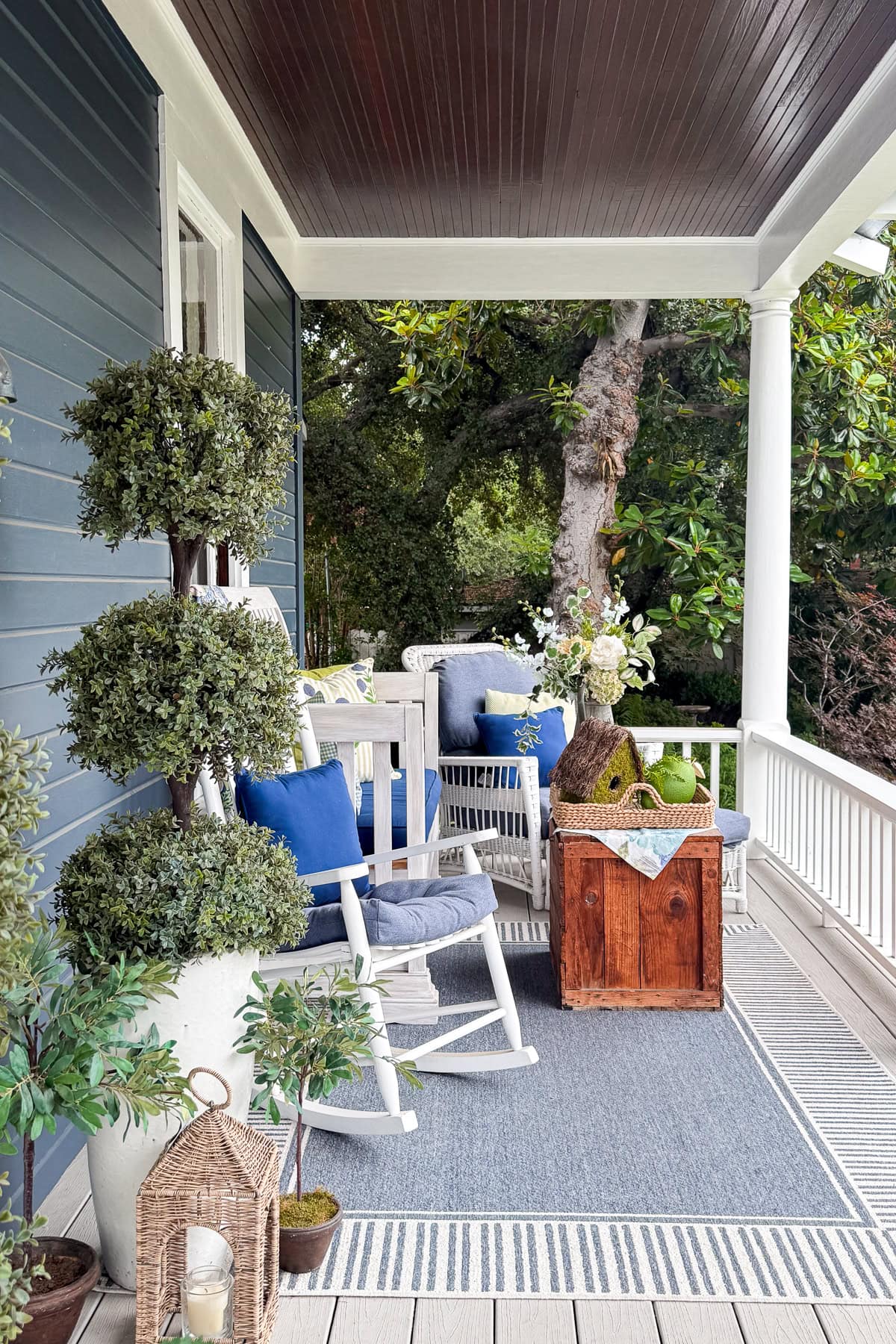 A cozy porch with a rocking chair, blue and white cushions, potted plants, a small wooden table with a basket, and a striped rug. The porch has white railings and overlooks green trees.