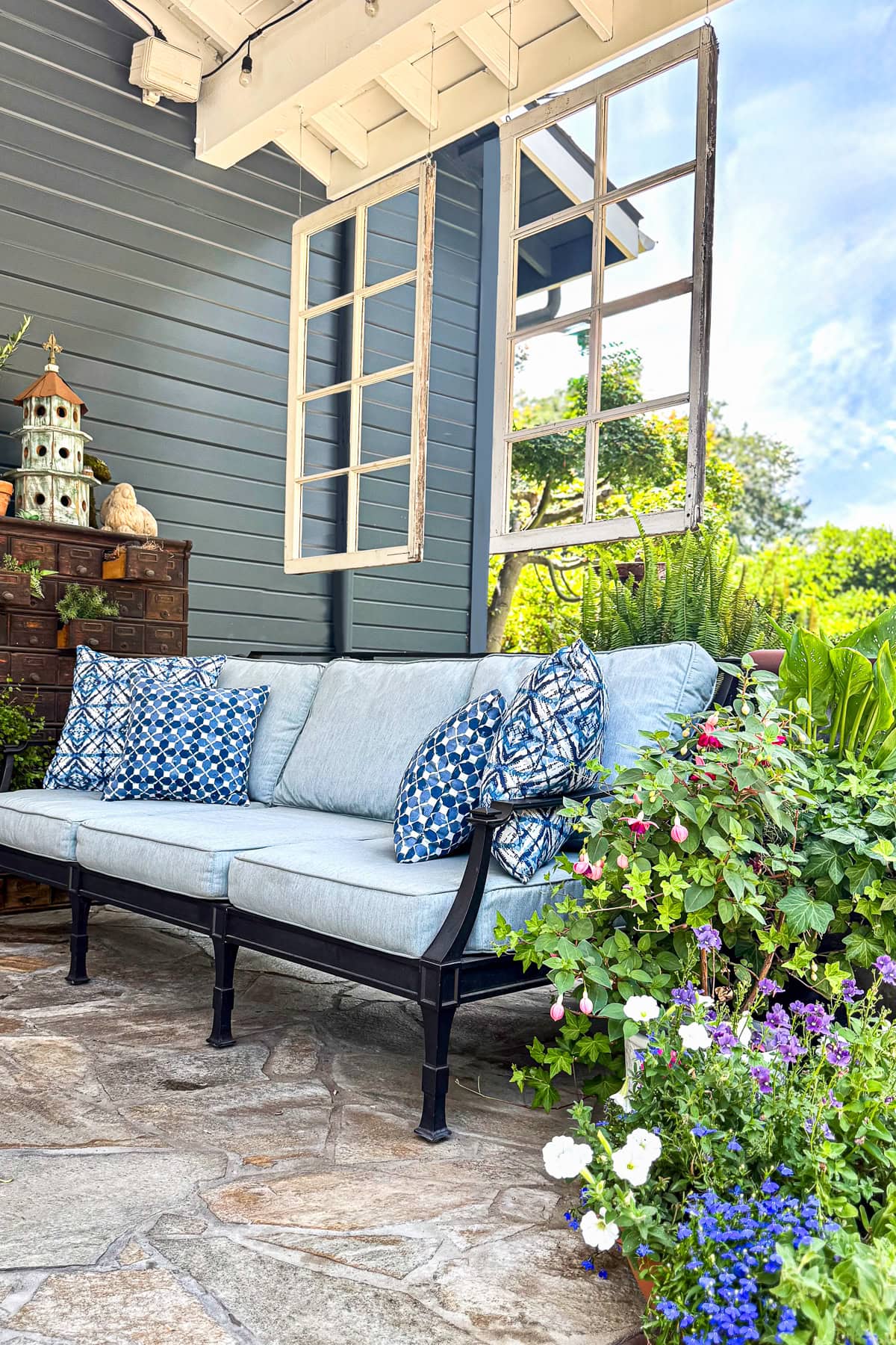 A cozy patio with a light blue cushioned sofa, blue pattern pillows, and potted flowers. Hanging window frames add charm. Lush greenery and blue sky are visible in the background.