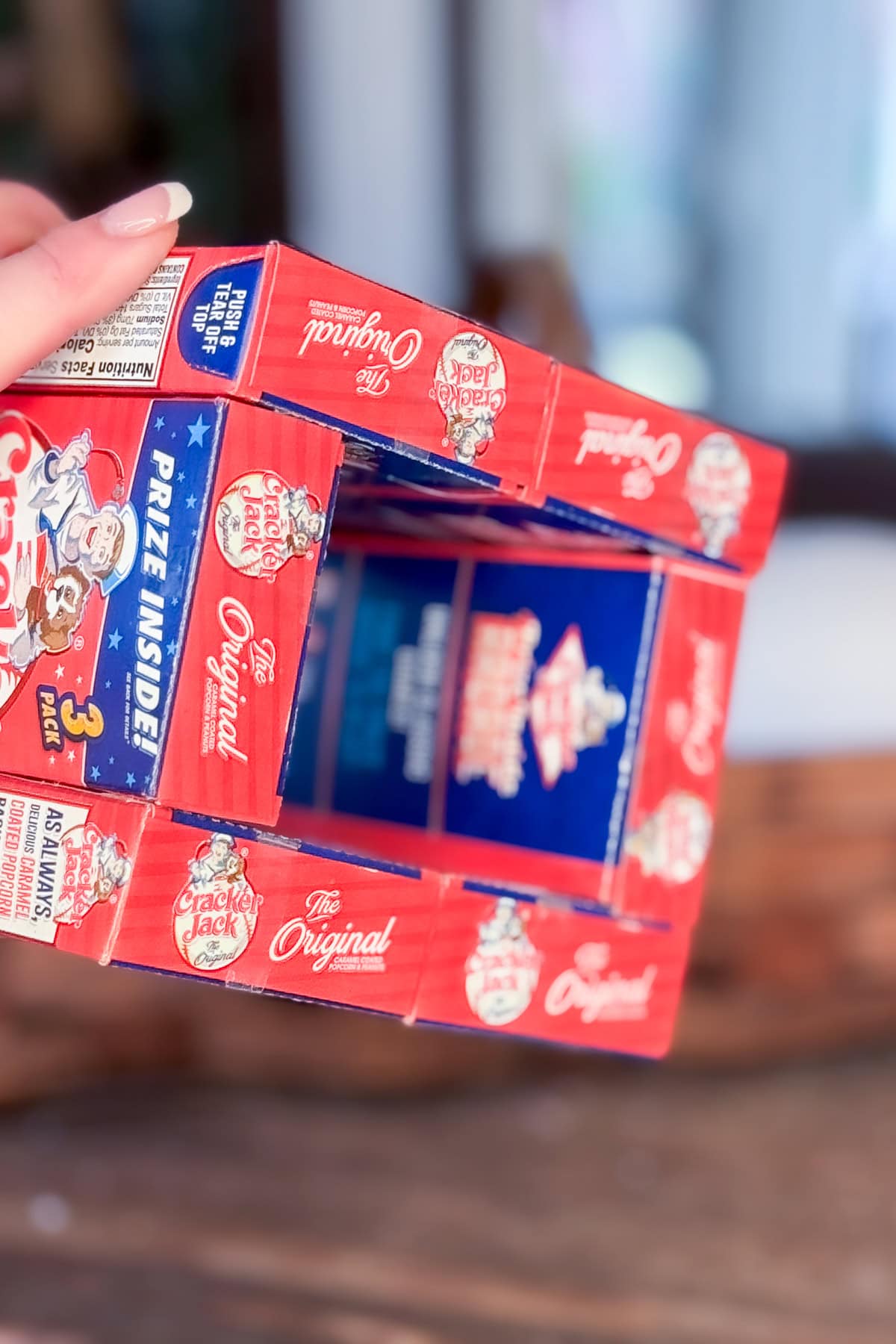 A hand holds a rectangular structure made from stacked Cracker Jack boxes, arranged with open ends facing forward. The boxes are red and blue, and the background is blurred.