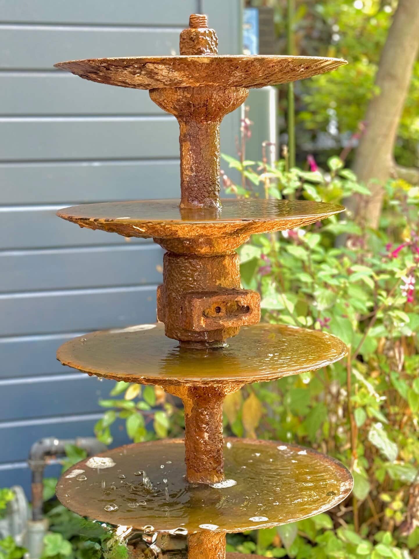 A close-up of a rusty, tiered metal water fountain with water flowing over each level, set outdoors in a garden with greenery and flowers in the background.