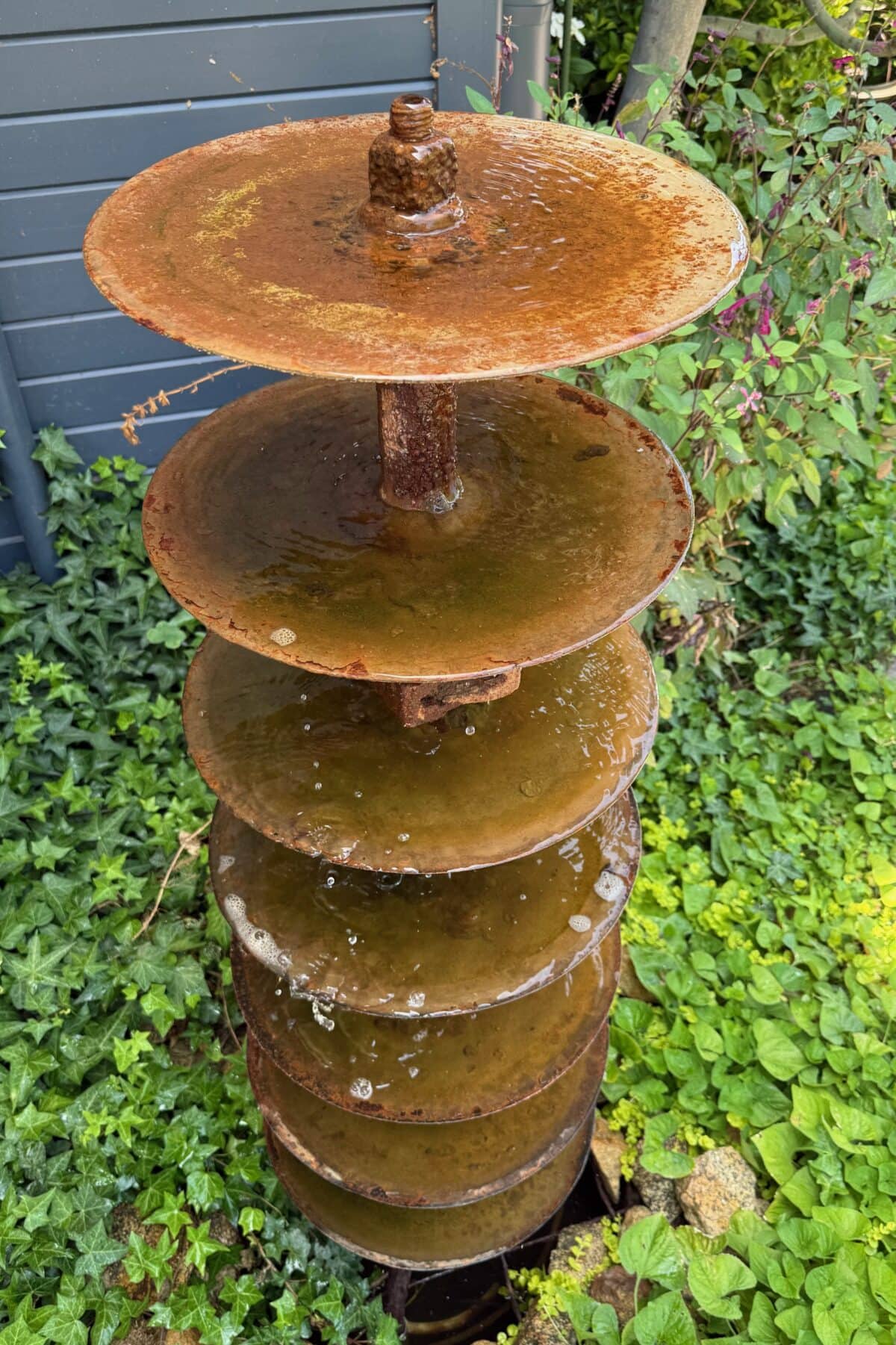 A rusty metal fountain made of stacked circular discs with water trickling down, surrounded by green leafy plants and ivy in a garden setting.
