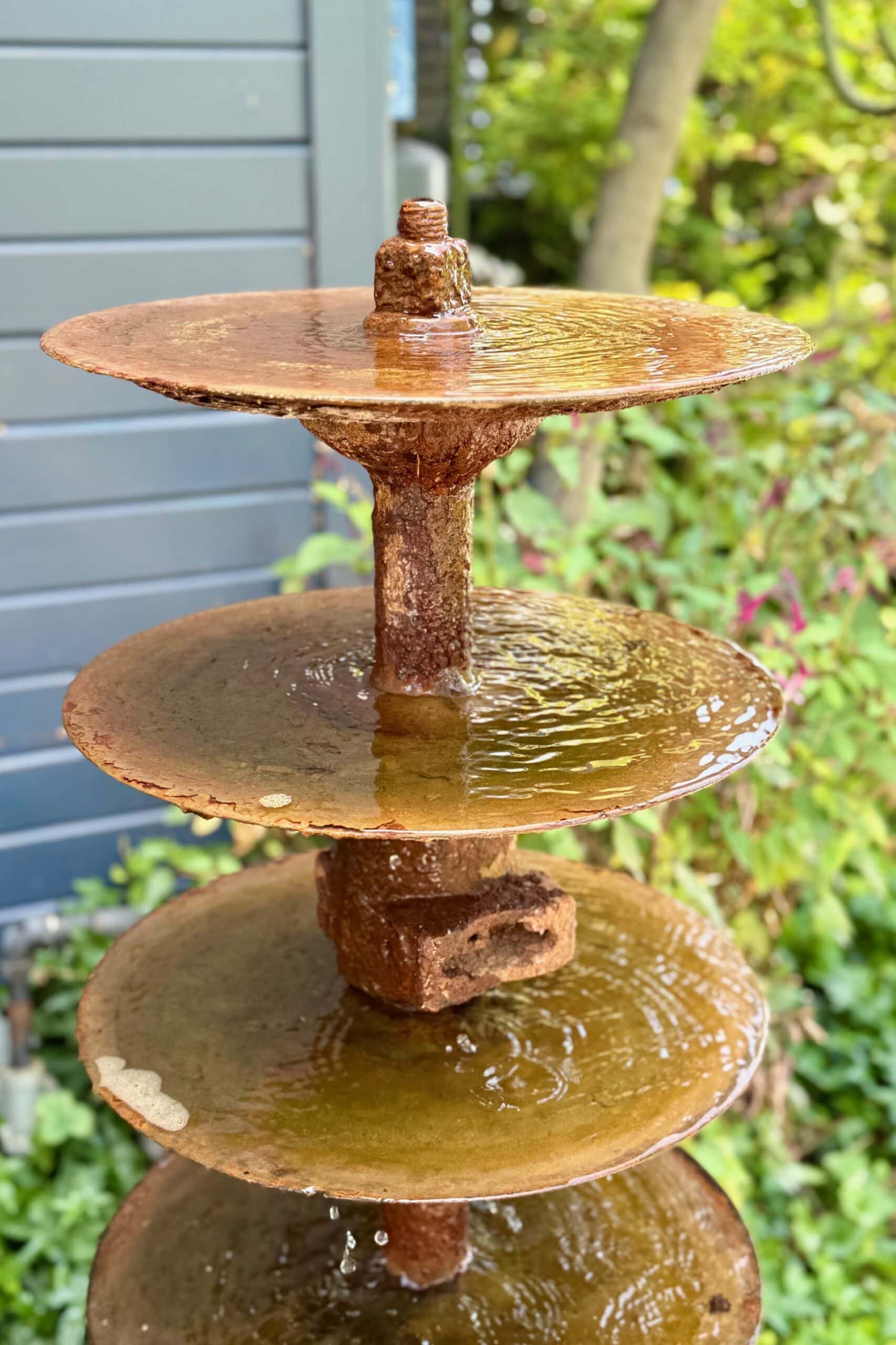 A close-up of a rustic, multi-tiered water fountain with circular, rusty metal plates, set outdoors with green foliage and a modern gray wall in the background. Water gently cascades from one tier to the next.