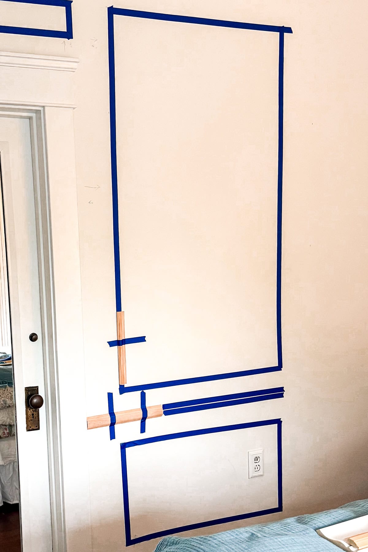 A white wall with two rectangles outlined in blue painter’s tape, one large and one small, with brown tape intersecting the lower rectangle. A doorframe and a visible electrical outlet are also present.