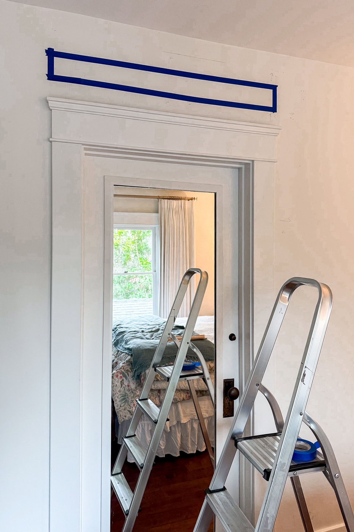 Two metal step ladders stand in front of a white door inside a room. Blue painter’s tape marks rectangles above the door on the wall, and a bed with floral bedding is visible through the doorway.