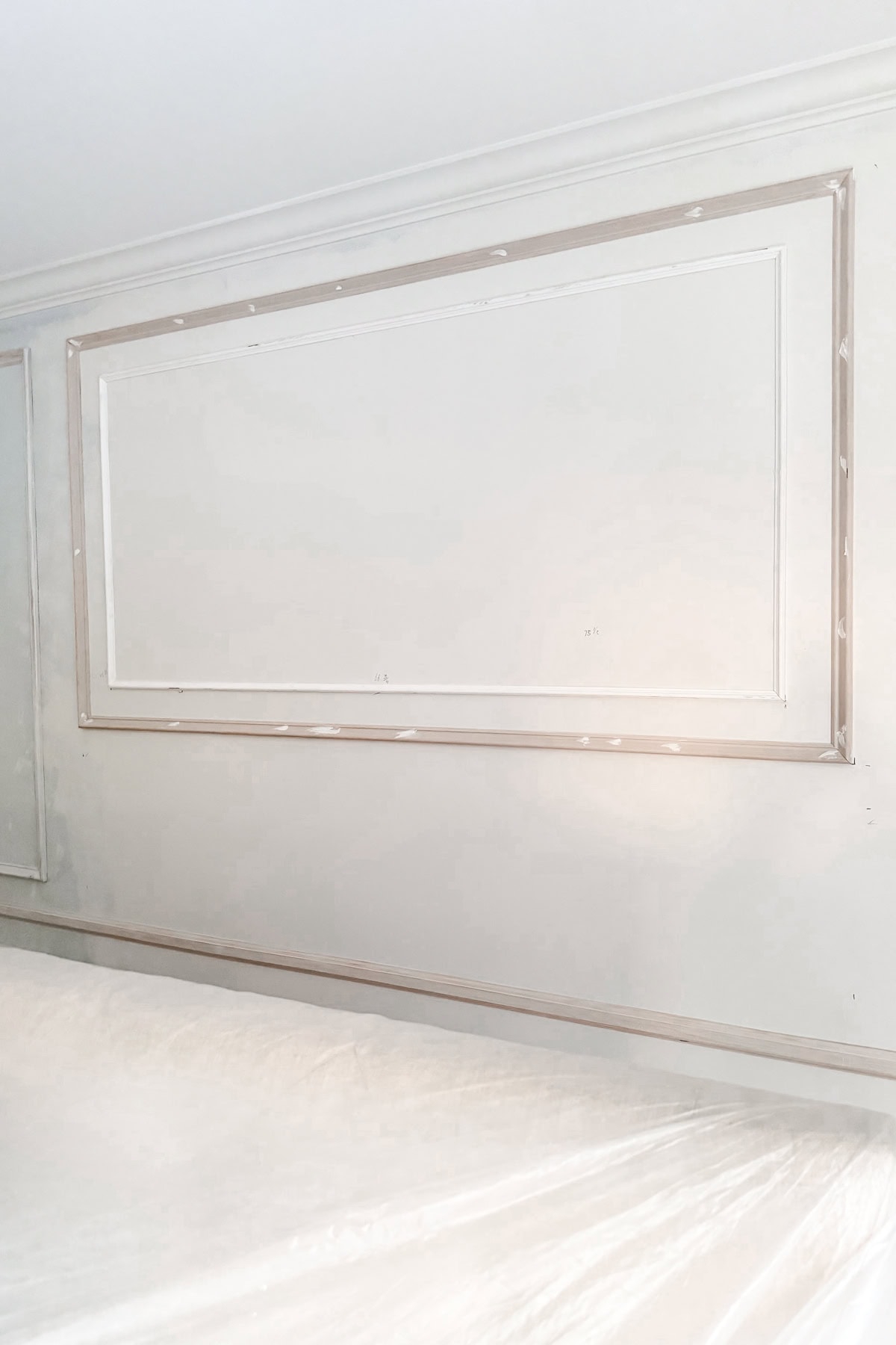 A white wall with rectangular DIY wall moldings in progress; some trim pieces are newly installed and patched. The lower portion of the image shows plastic covering furniture or flooring for protection.
