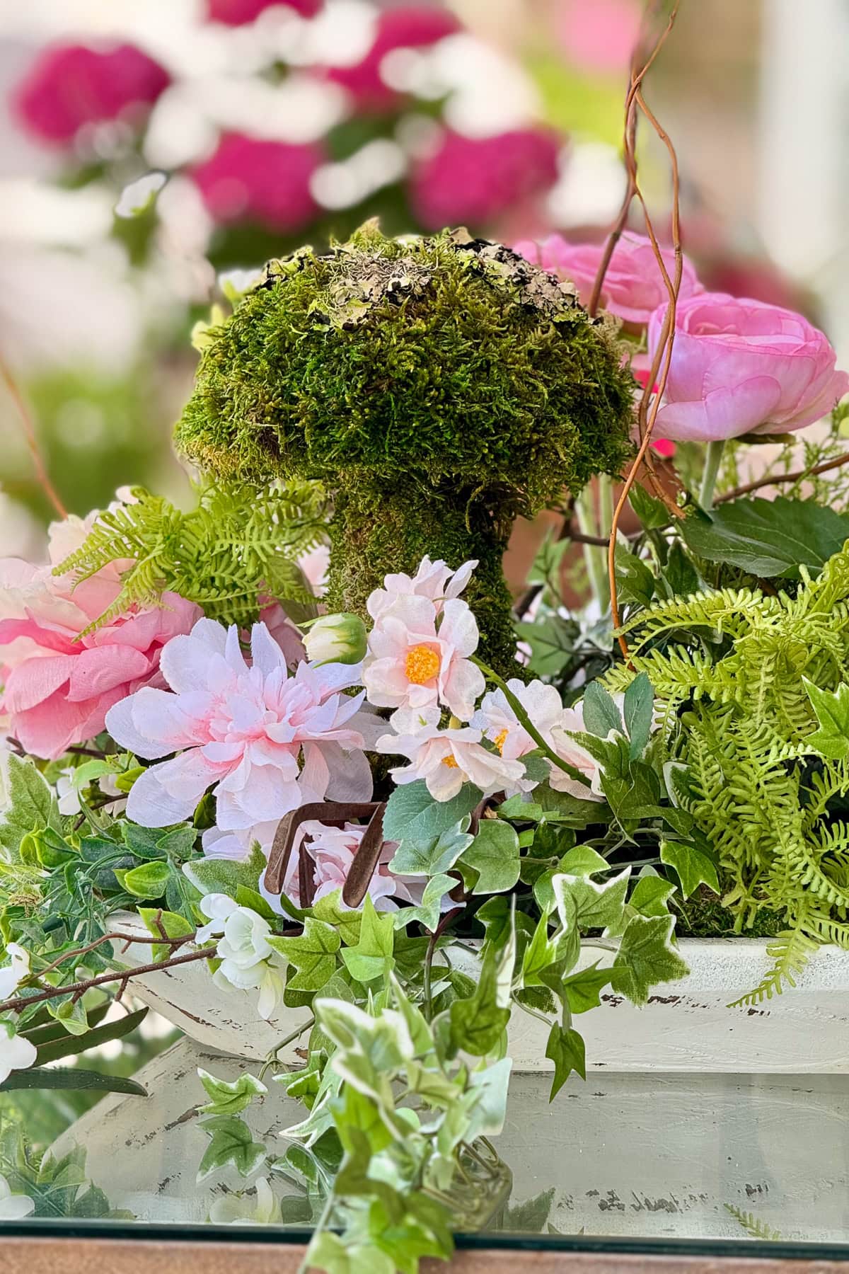 A decorative arrangement featuring a moss-covered mushroom, pink and white artificial flowers, ferns, and green ivy in a white container, with blurred pink blooms in the background.