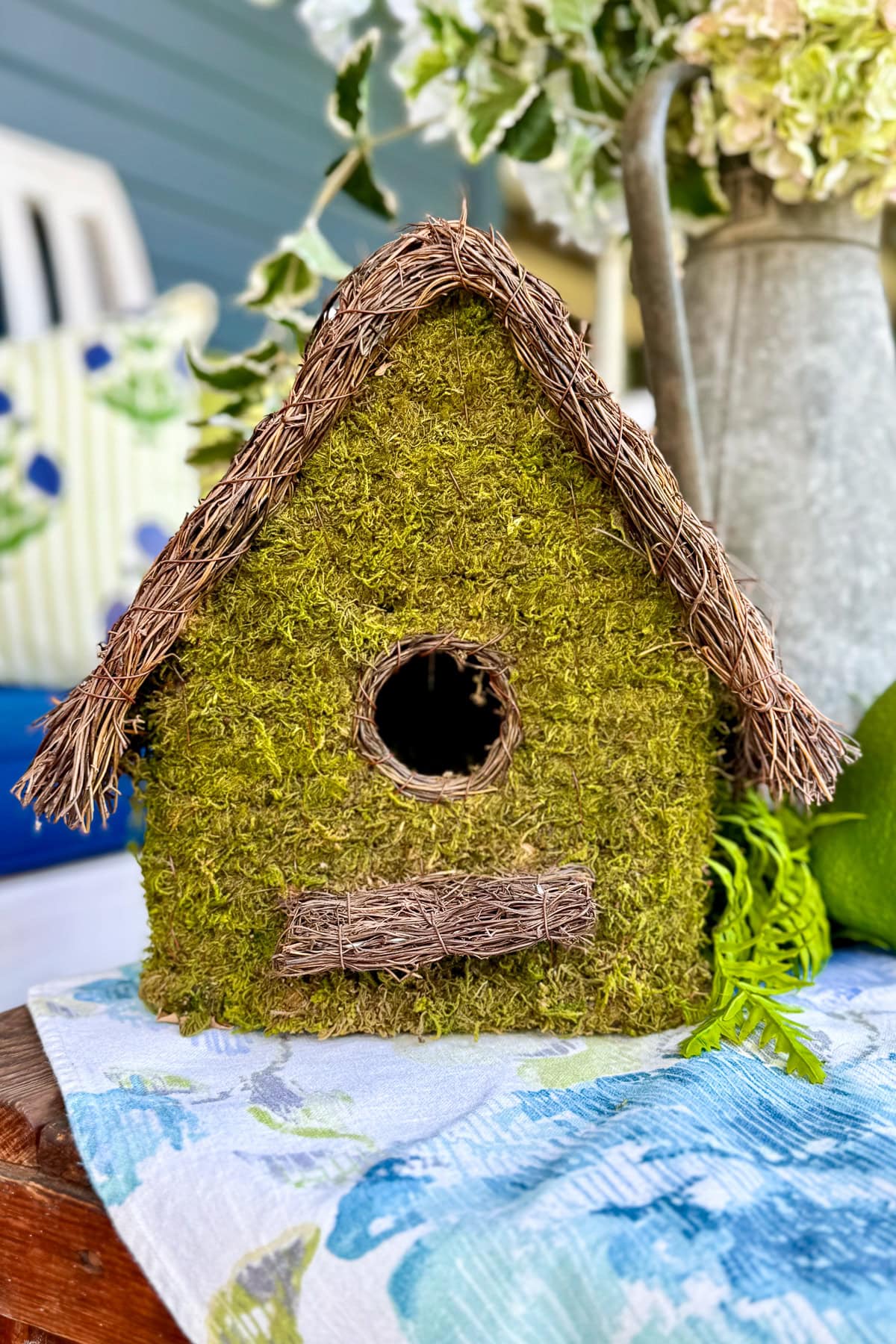 A small, rustic birdhouse covered in green moss with a circular entrance, perched on a wooden surface and surrounded by flowers and greenery.