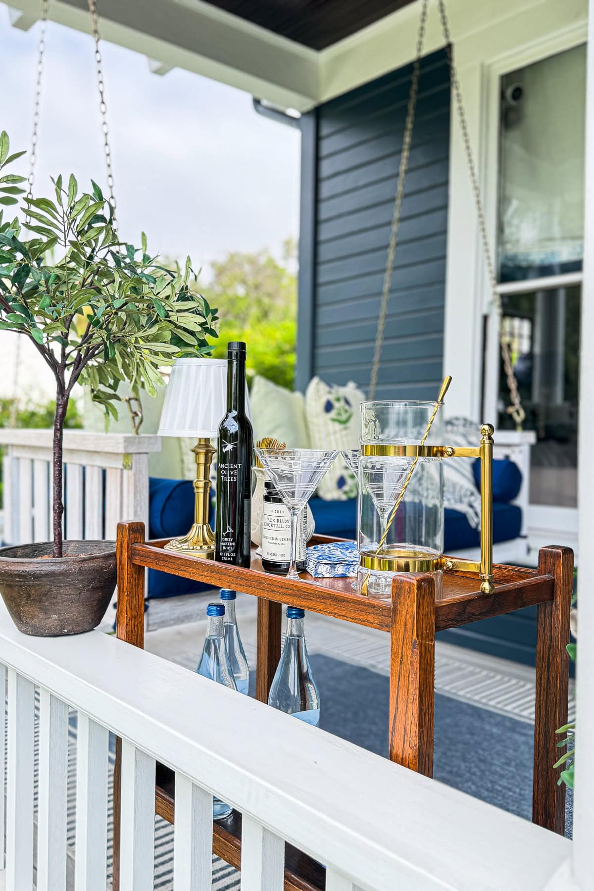 A wooden bar cart on a porch displays bottles, glasses, a lamp, and a potted plant, with a blue house and white railing in the background. Two bottles of water are stored on the cart’s lower shelf.