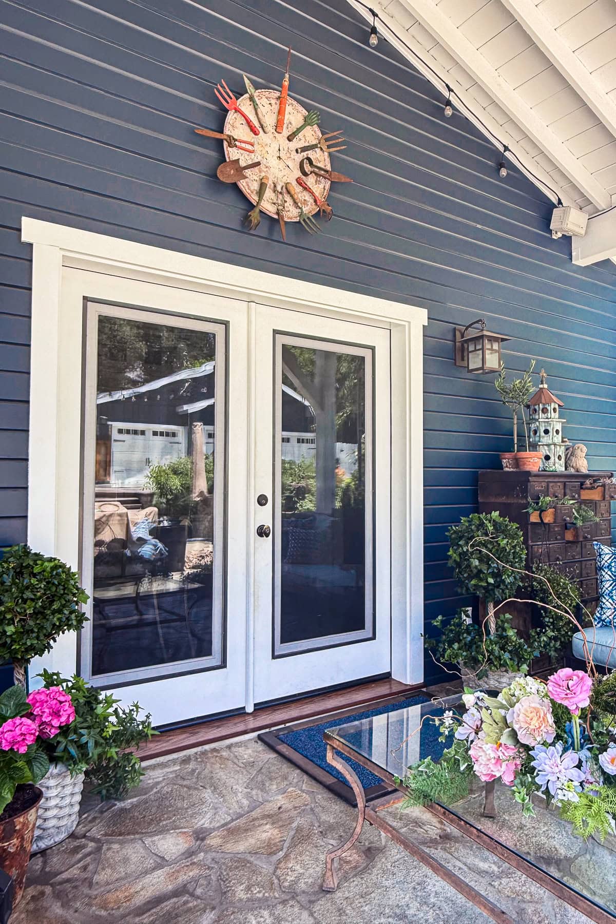 A pair of white French doors with glass panes open onto a porch with blue siding. Decorative plants, flowers, and a birdhouse sit nearby. A whimsical clock made from garden tools hangs above the door.