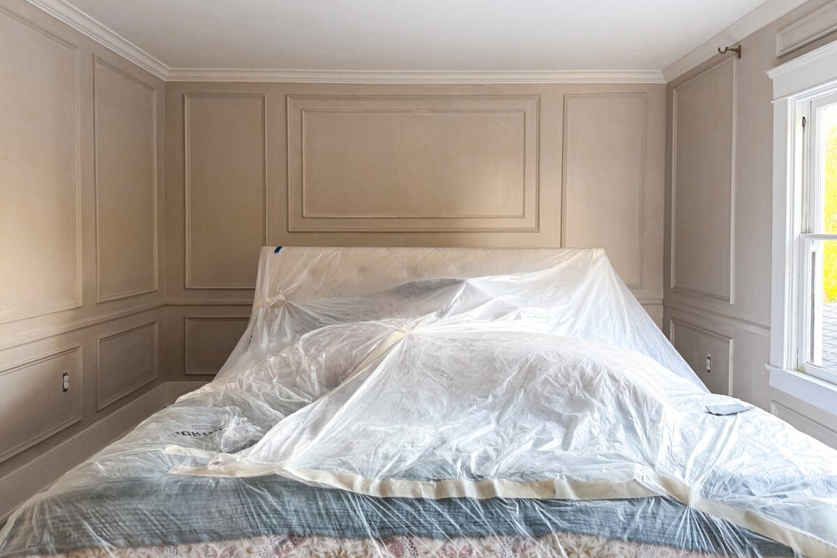 A bedroom with tan paneled walls featuring DIY wall moldings and a large bed covered completely with clear plastic sheeting, likely for protection during painting or renovation. Sunlight enters through a window on the right.