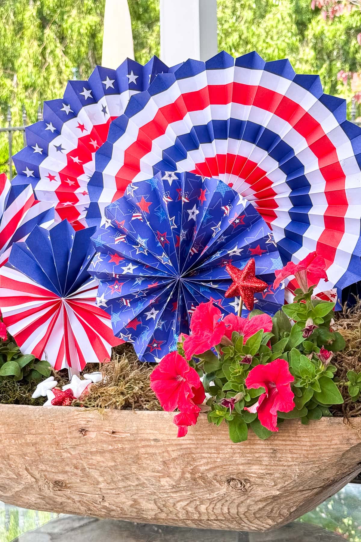 A wooden planter with bright pink flowers and festive red, white, and blue paper fans decorated with stars and stripes, creating a patriotic display.