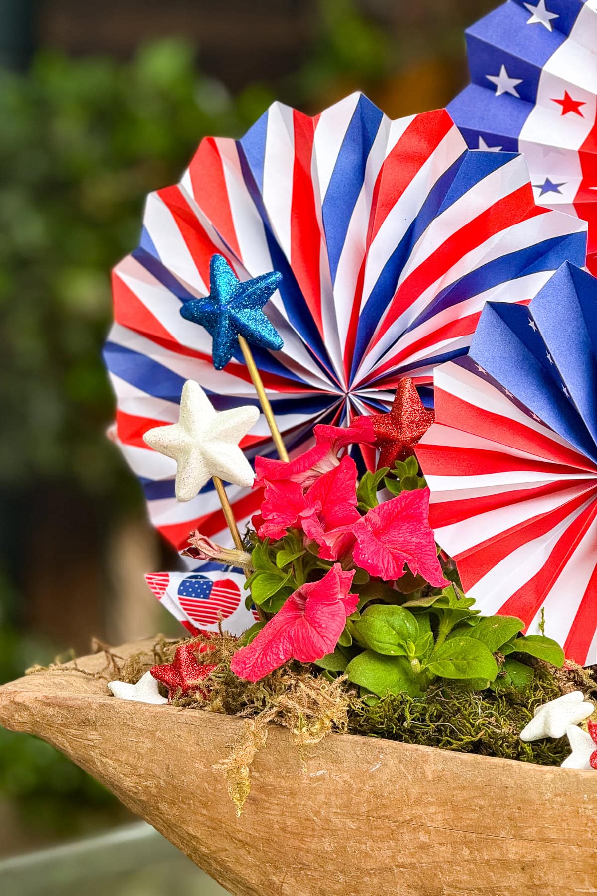 A festive display features red, white, and blue paper fans, glittery star decorations, and pink flowers in a rustic planter, evoking a patriotic theme.