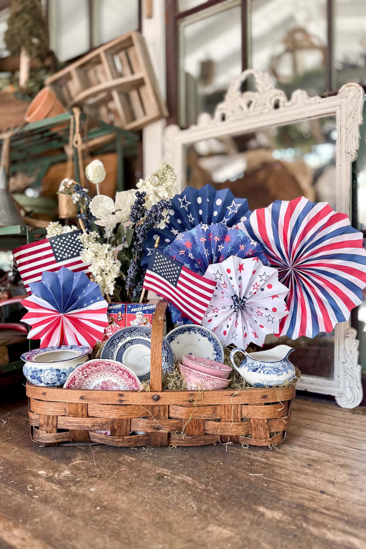 A wicker basket holds vintage red, white, and blue dishes, American flags, festive paper pinwheels, and white flowers, set on a rustic wooden table with a large mirror and farmhouse decor in the background.