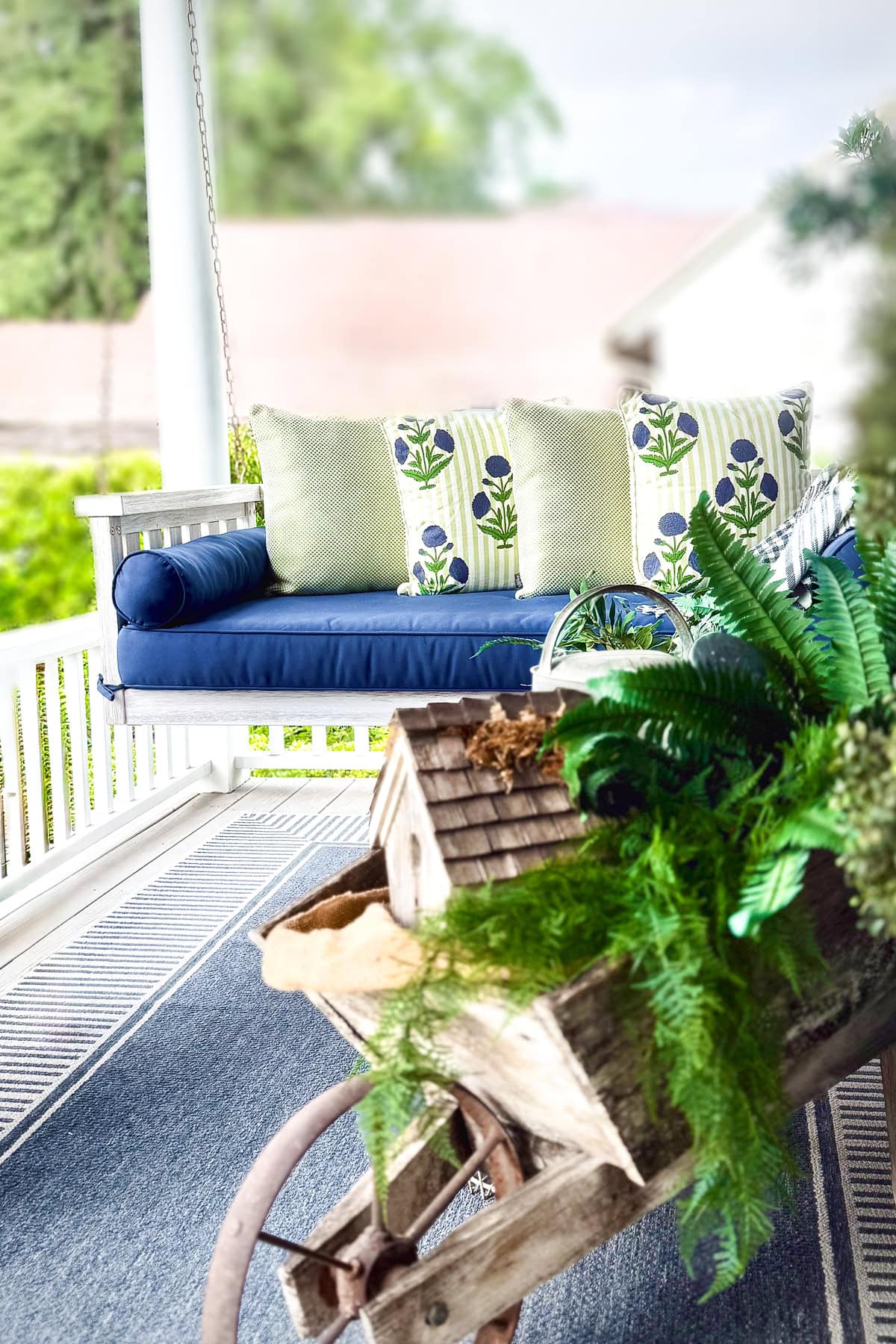 A porch swing with blue cushions and floral pillows sits on a blue rug, behind a rustic wooden wheelbarrow planter filled with green ferns and plants. Trees and a house roof are visible in the blurred background.