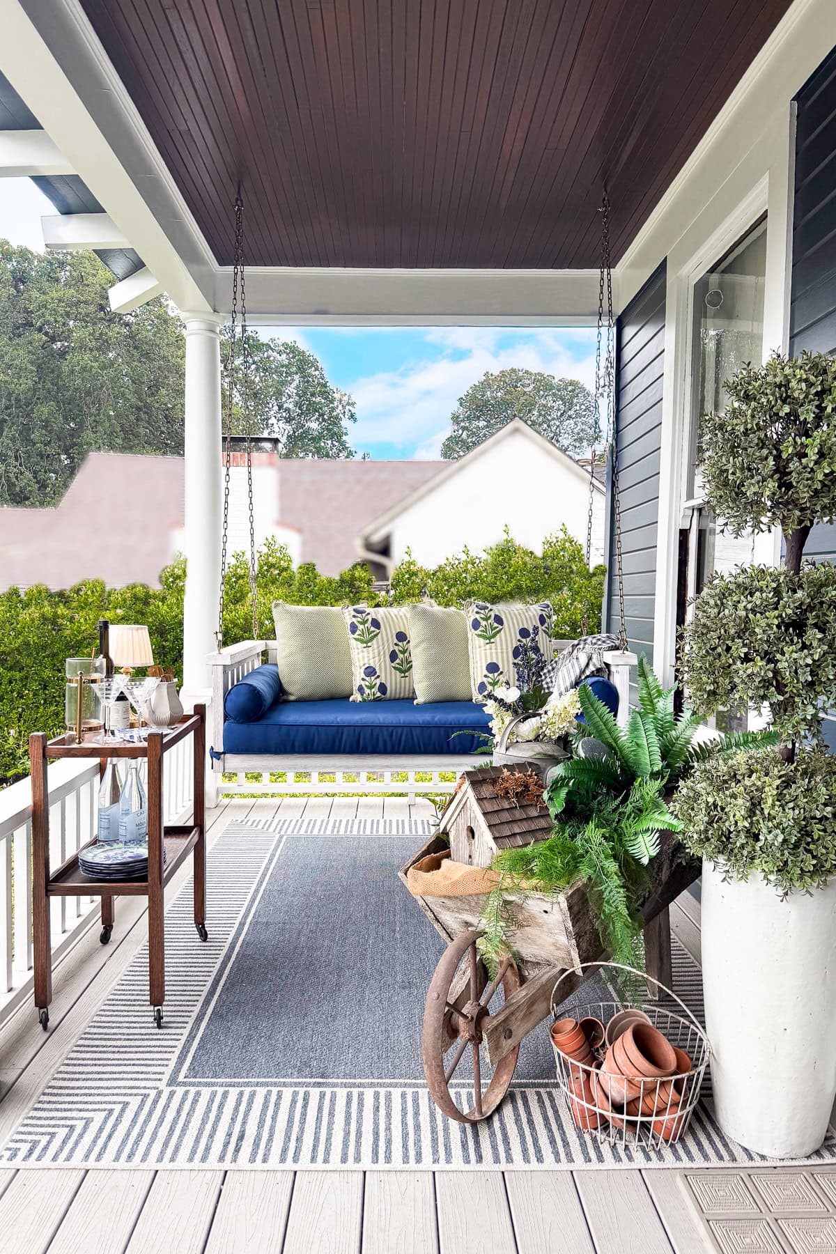 A cozy front porch with a blue cushioned swing, striped rug, potted plants, a decorative wheelbarrow, gardening tools, and a small drinks cart, all set under a covered roof with white columns.