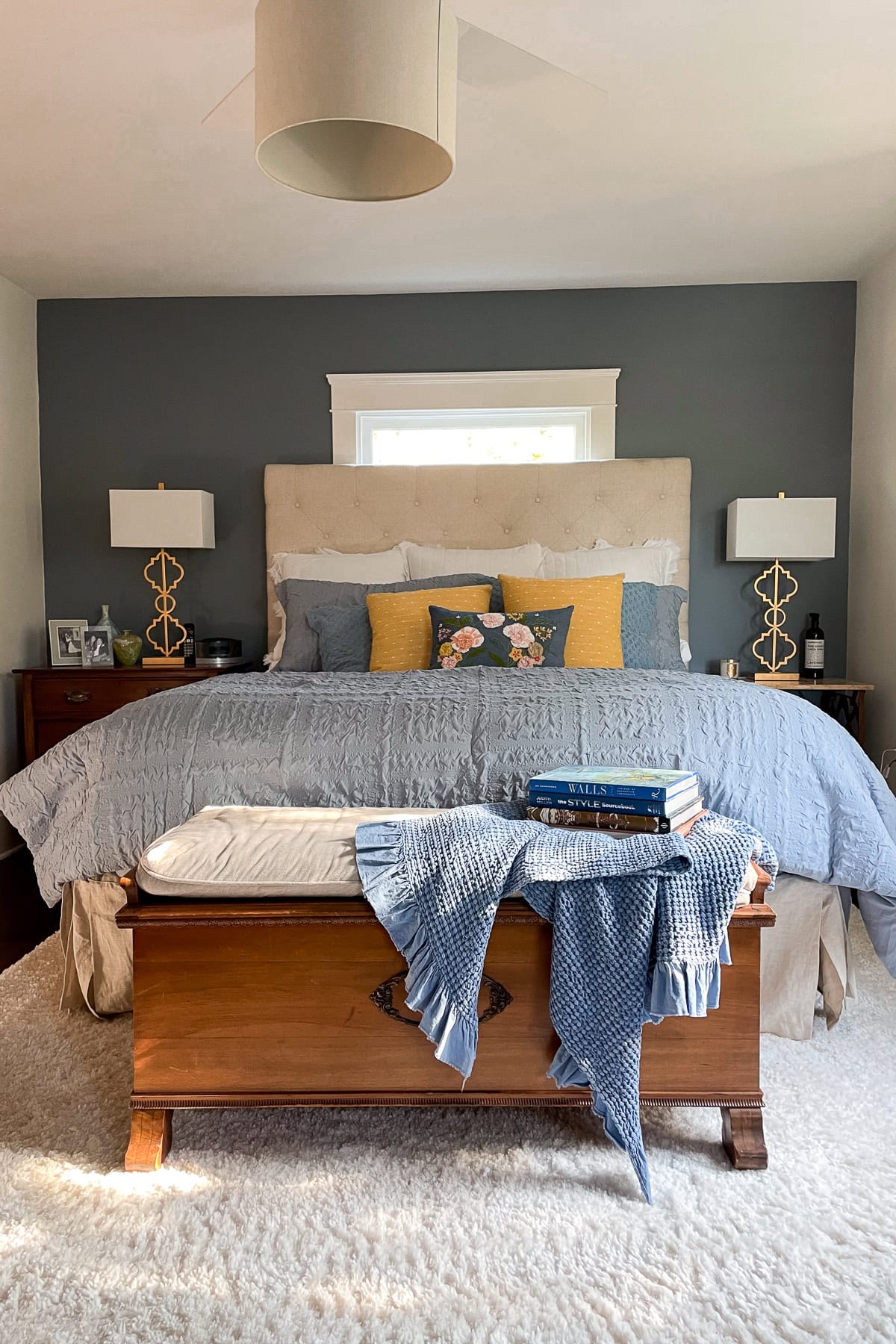 A cozy bedroom features DIY wall moldings, a neatly made bed with blue and yellow bedding, decorative pillows, and a beige headboard. A wooden bench with books and a blue blanket sits at the foot of the bed; matching lamps are on both nightstands. This is our bedroom before painting it Benjamin Moore Smokey Taupe. 